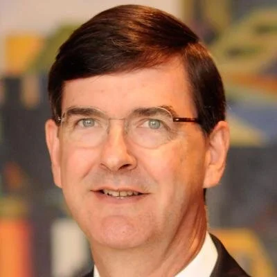 A middle-aged man with short dark brown hair, glasses, and a slight smile, wearing a suit, with a colorful blurred background.