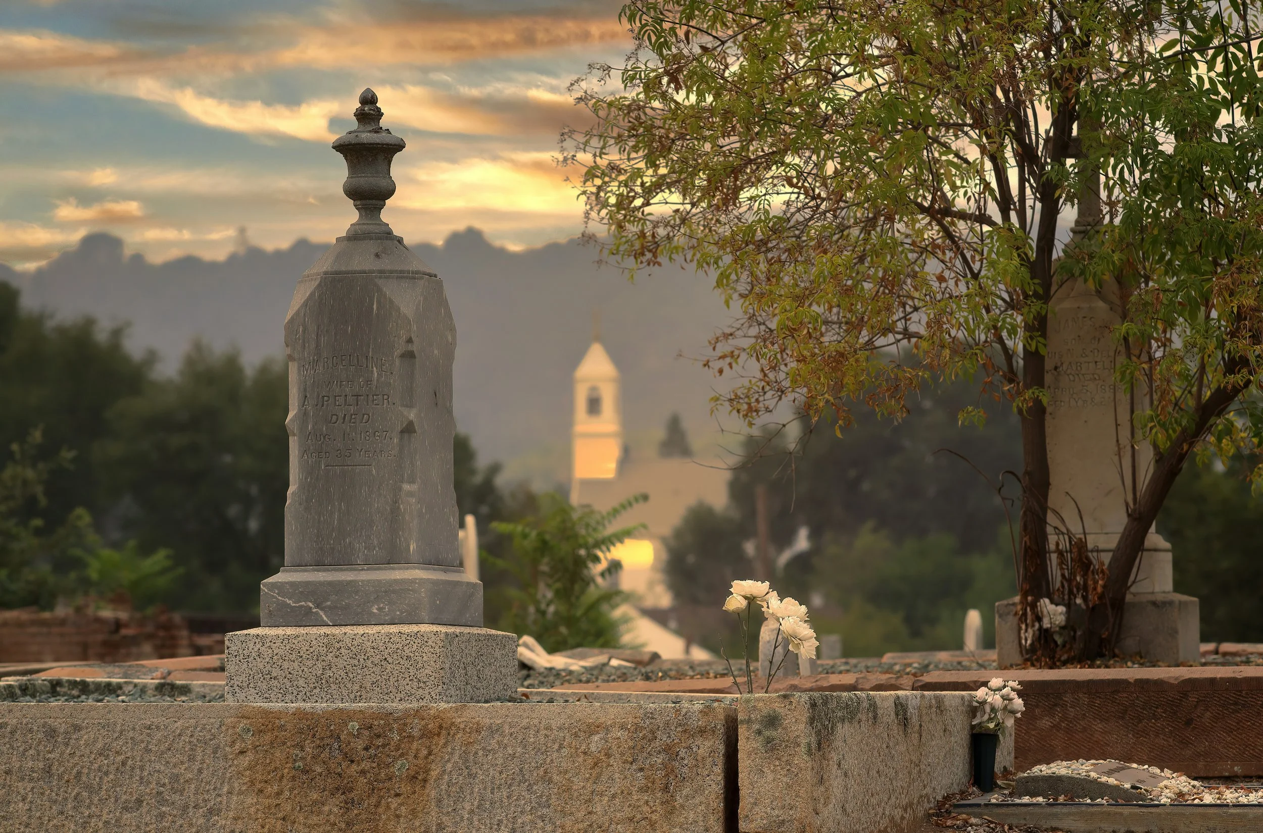 A cemetery scene at sunset with a large gray tombstone in the foreground, unreadable inscriptions, and a small bouquet of white flowers nearby. There are other tombstones partially obscured by trees, and a church steeple is visible in the distance against a colorful sky with clouds.