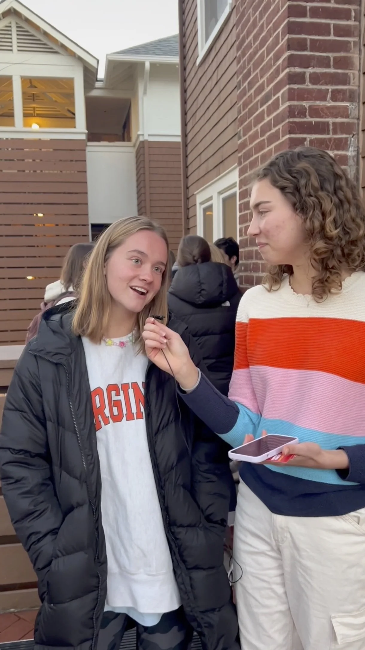 Two young women having a conversation outside near a brick building, with other people in the background. One woman is holding a smartphone and appears to be interviewing or asking questions to the other woman.