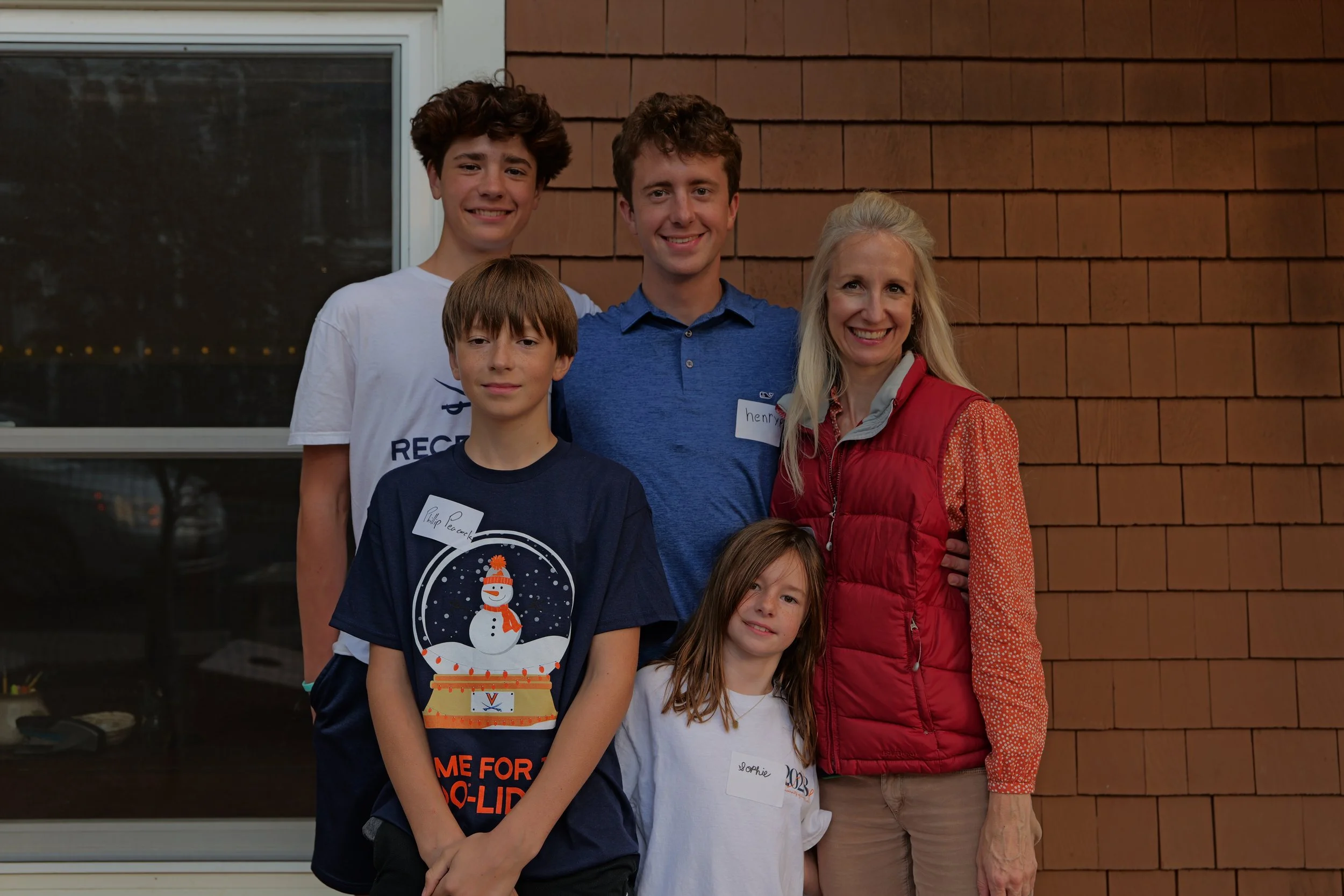 A group of six people, four children and two adults, standing together outside near a brick and window wall, smiling at the camera, with Christmas-themed attire on one child.