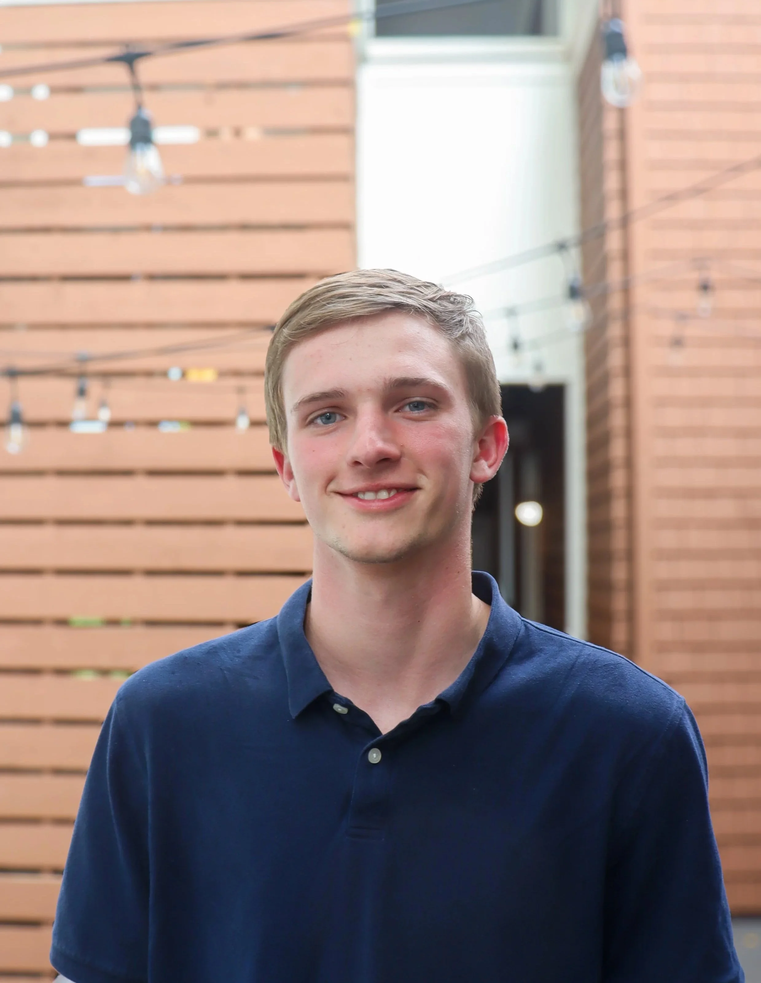 A young man with blonde hair and blue eyes standing outdoors in front of a modern wooden building with string lights hanging above him, smiling at the camera.