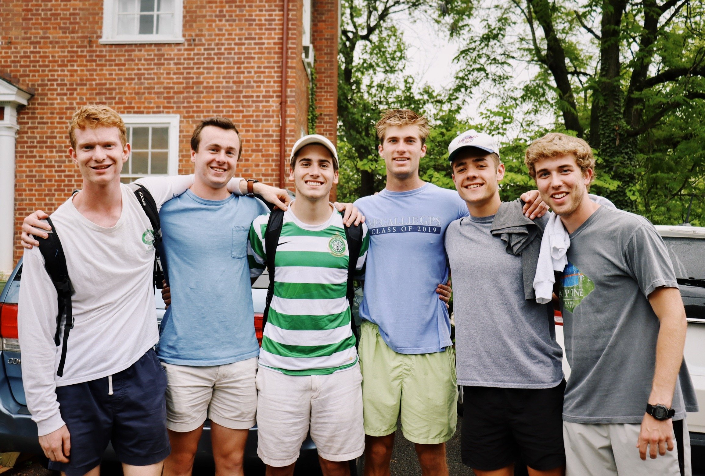 Six young men standing outside in front of a brick building and trees, smiling, with arms around each other.