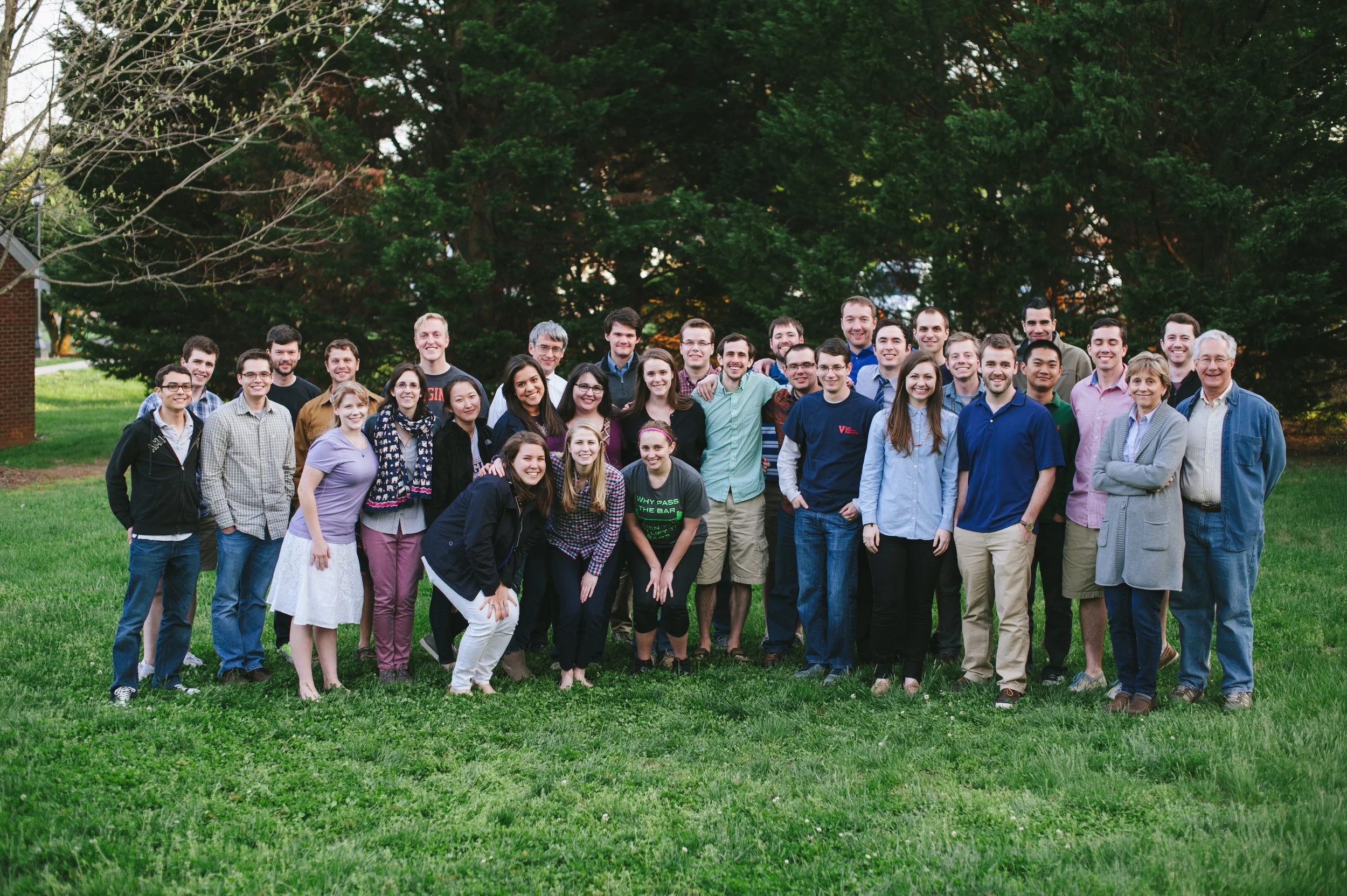 A large group of diverse people posing outdoors on a grassy area with trees in the background.