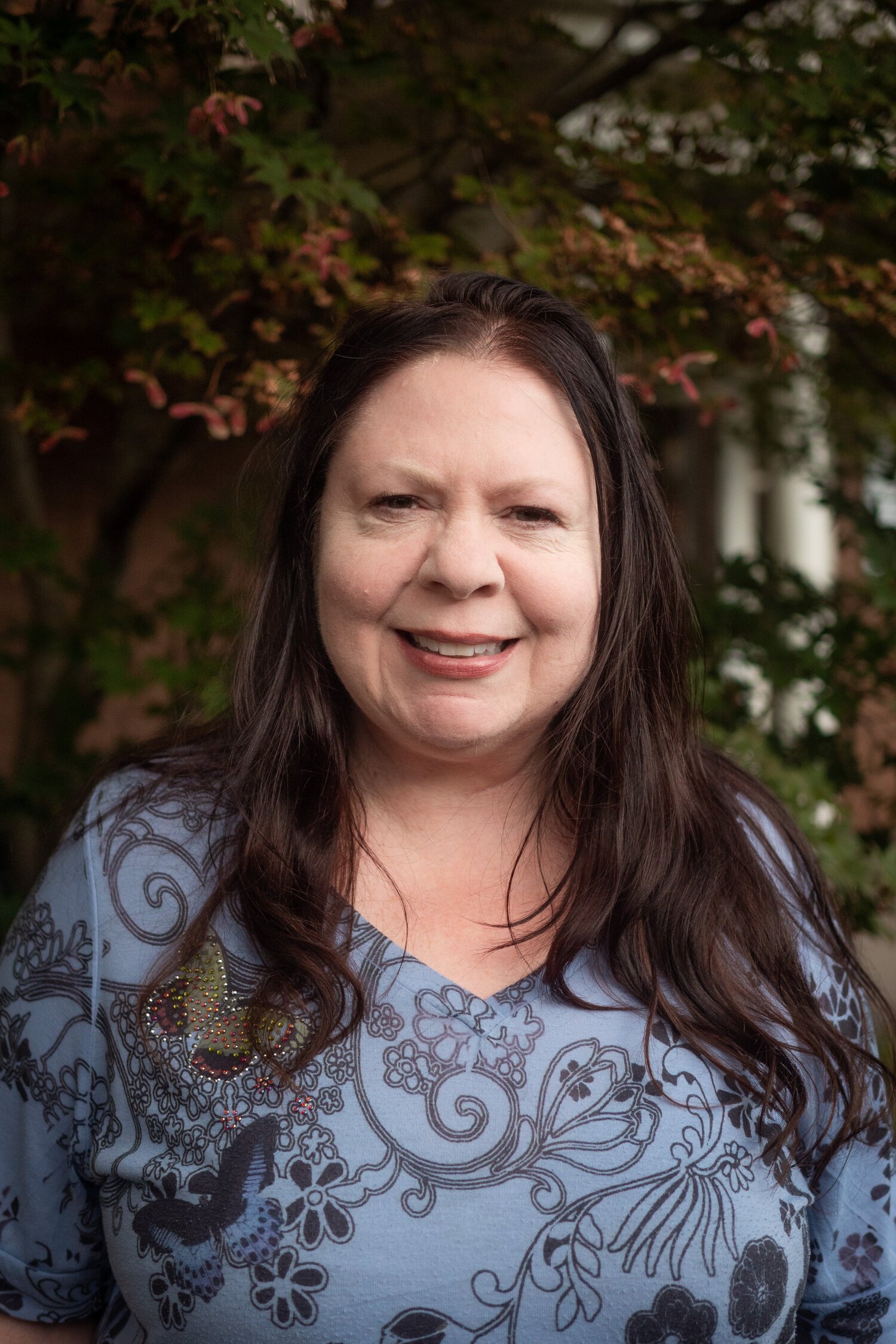 A smiling woman with dark hair and braces, wearing a patterned top, standing outdoors with green foliage in the background.