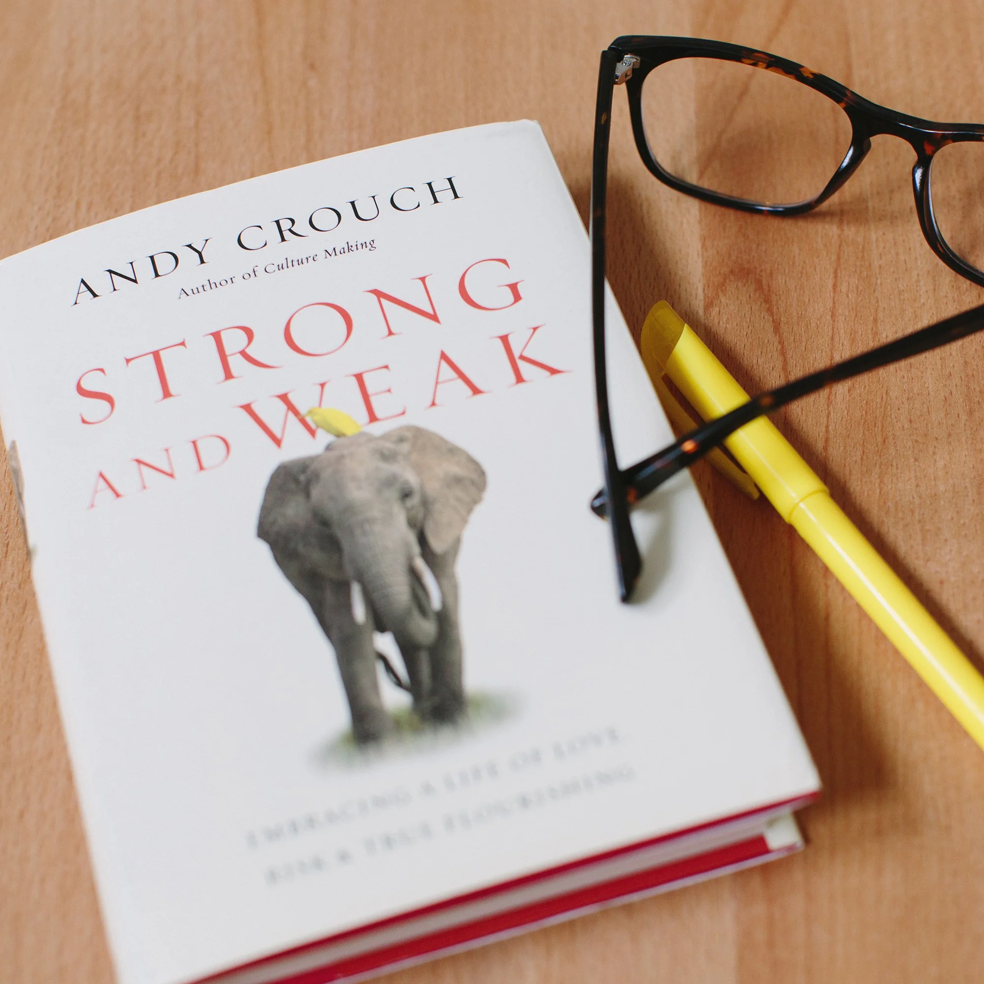 Book titled 'Strong and Weak' by Andy Crouch, with a photograph of an elephant on the cover. Glasses, a yellow highlighter, and a yellow pen are on a wooden surface beside the book.