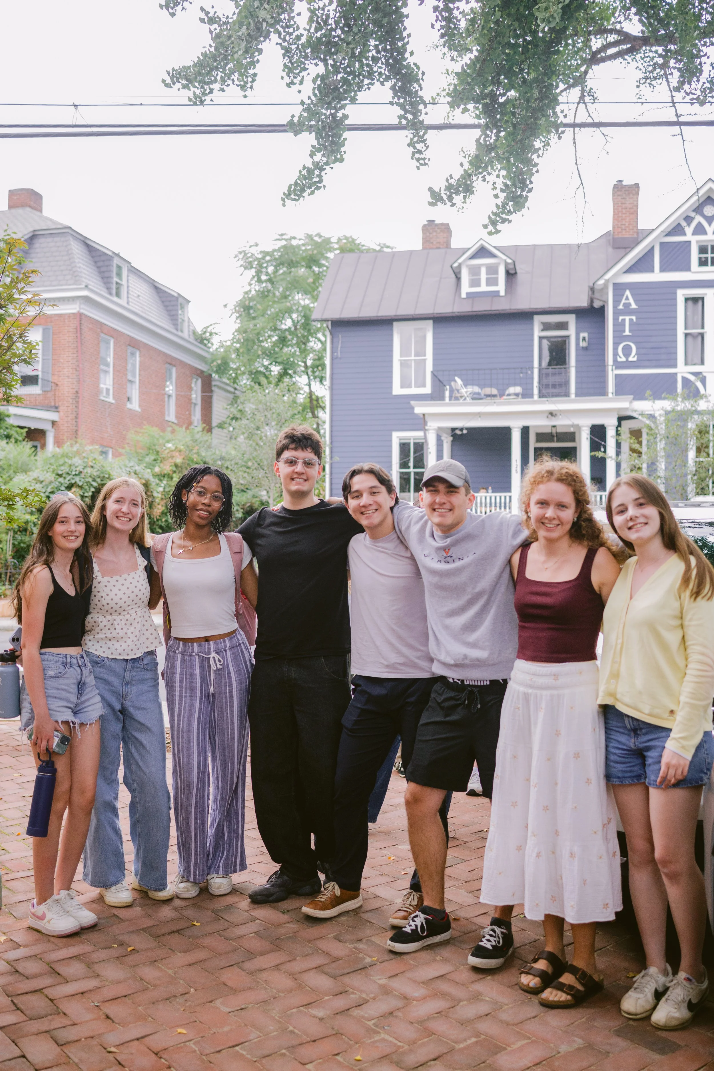 Group of eight young people standing on a brick sidewalk in front of colorful houses, smiling and posing with arms around each other.