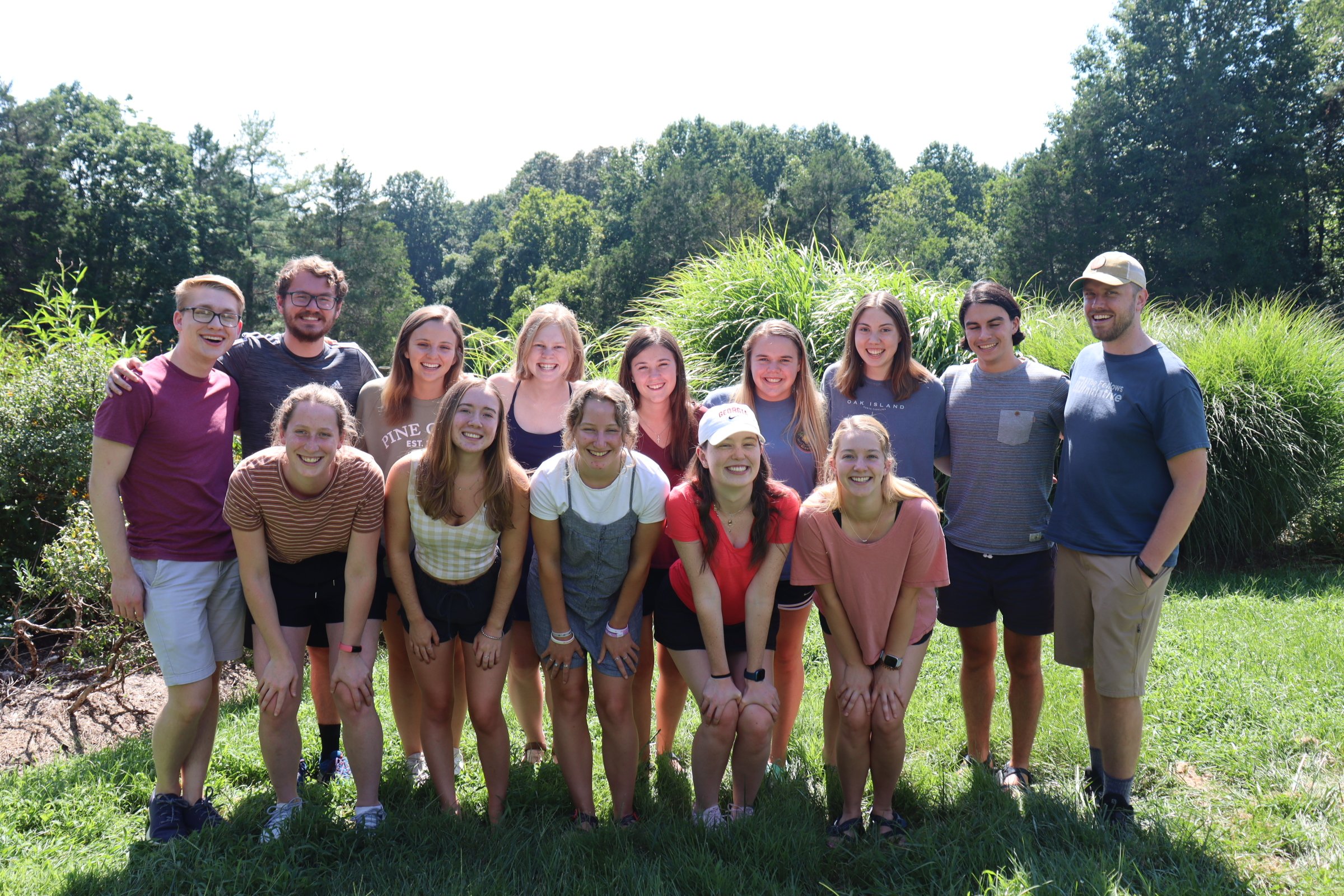 Group of 15 young adults outdoors on a sunny day, standing on grass with trees and bushes in the background, smiling and posing for the photo.