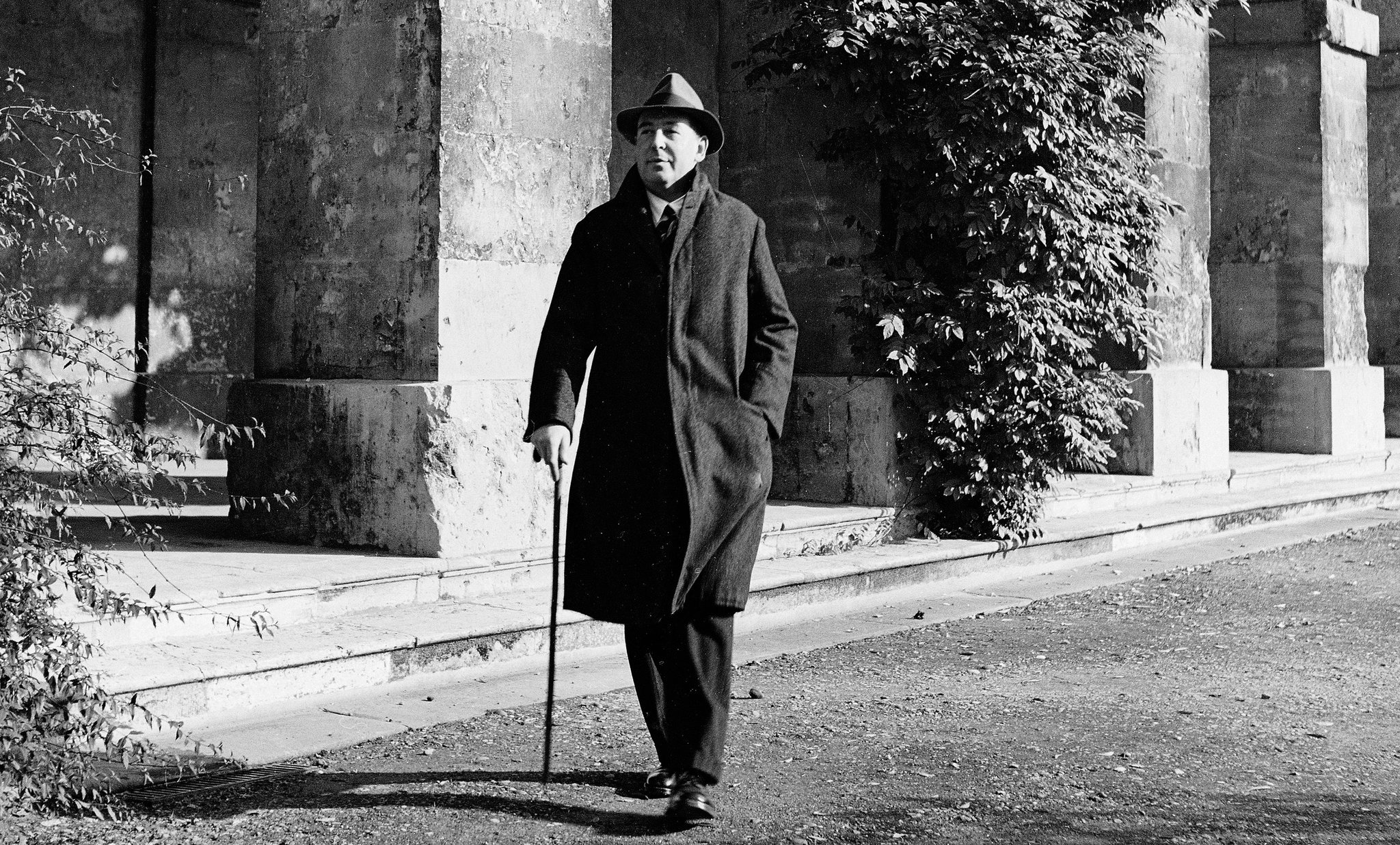 Black and white photo of a man wearing a long coat and hat, walking with a cane beside a stone building with columns and greenery.