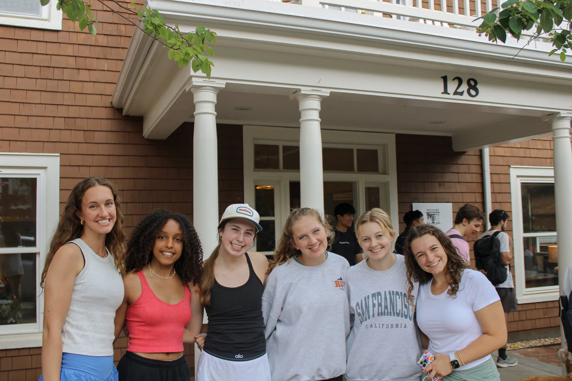 Six young women standing outside, smiling at the camera, in front of a house with the number 128, and other people in the background.