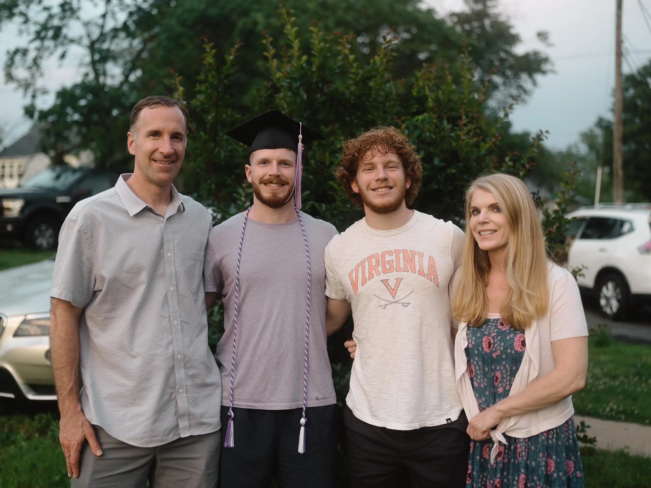 A family of four outdoors celebrating a graduation, with a young man wearing a cap and gown, and his parents standing beside him.