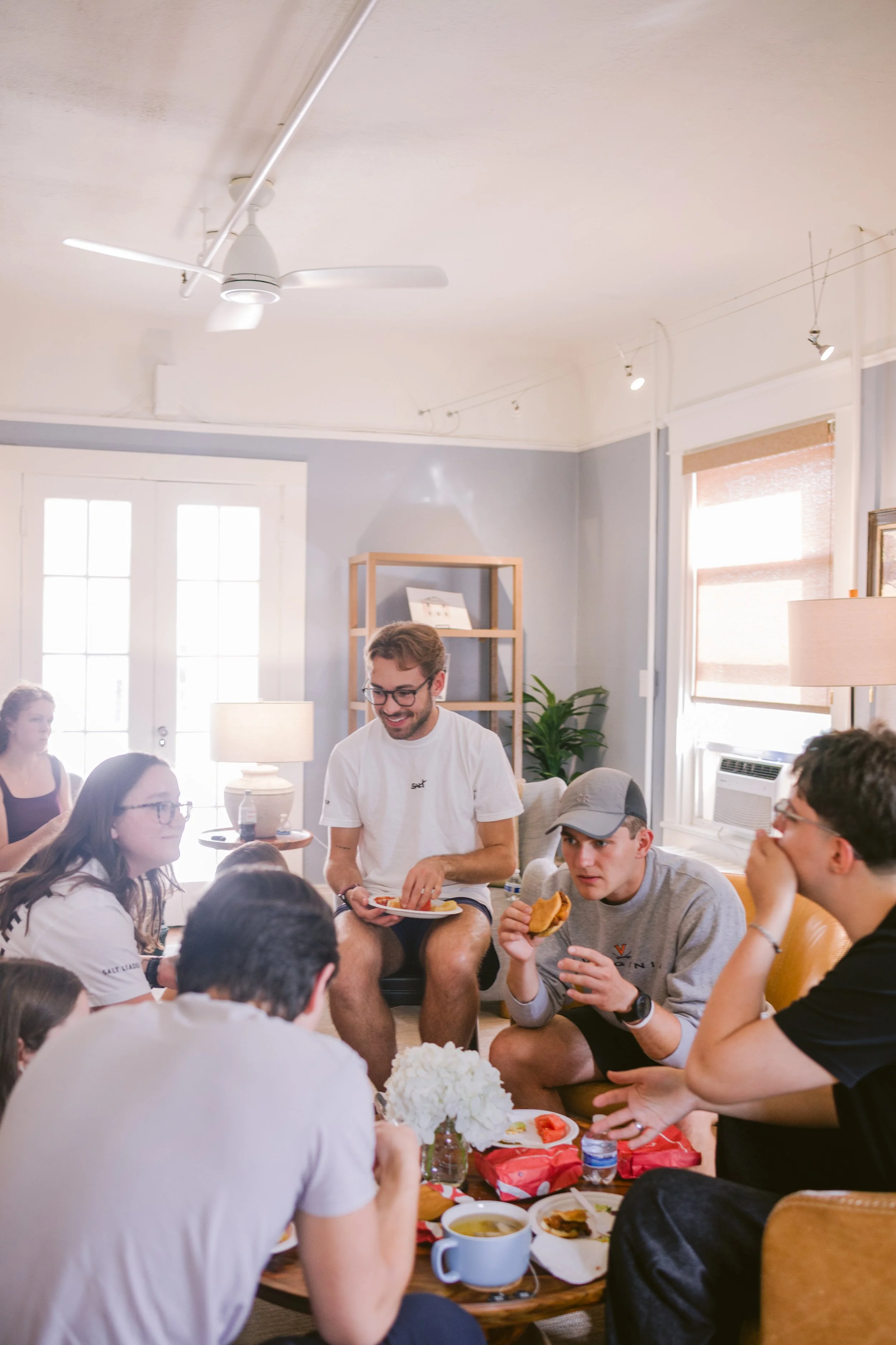 Group of young adults gathered around a table in a bright living room, enjoying food and conversation, with a ceiling fan and large windows in the background.