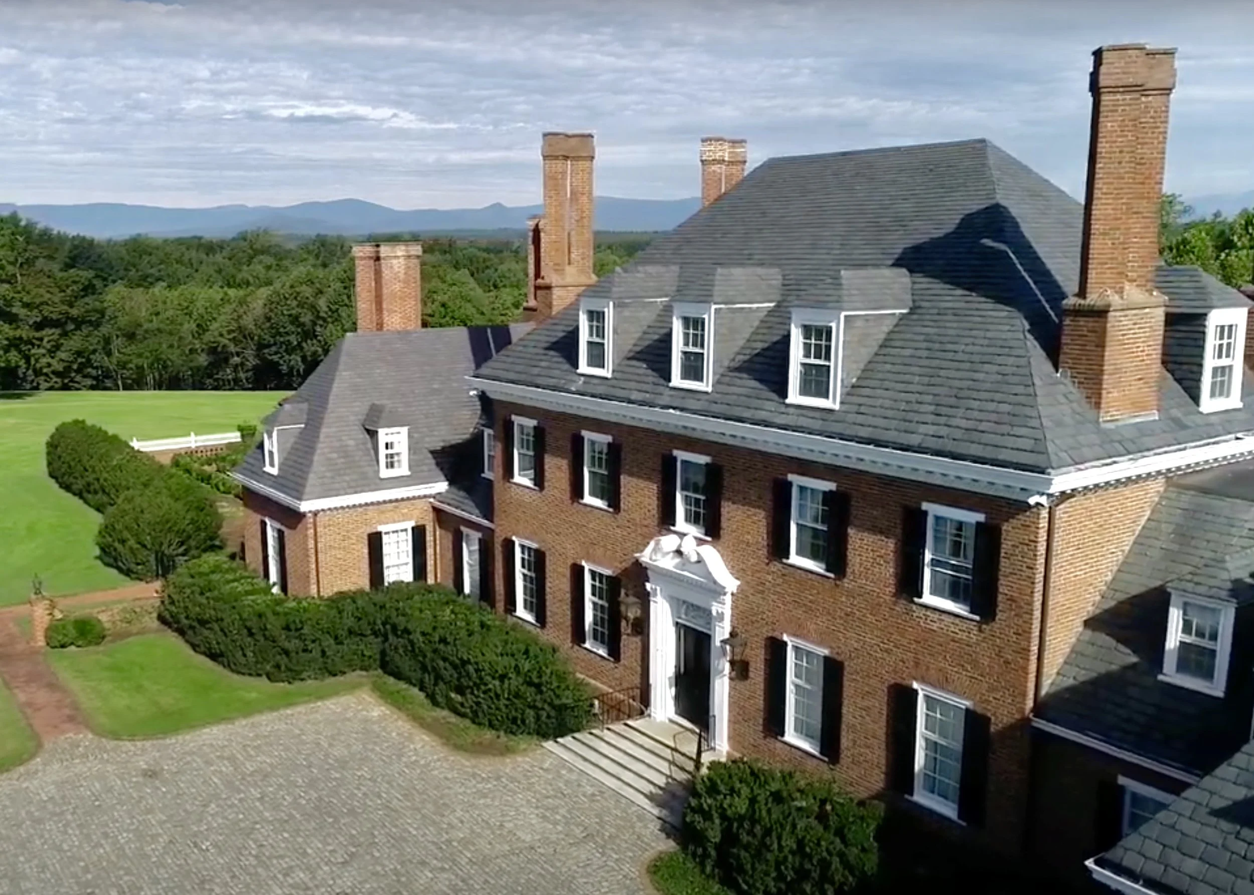 A large brick mansion with black shutters and multiple chimneys, surrounded by neatly trimmed hedges and a grassy lawn, with distant mountains and a partly cloudy sky in the background.
