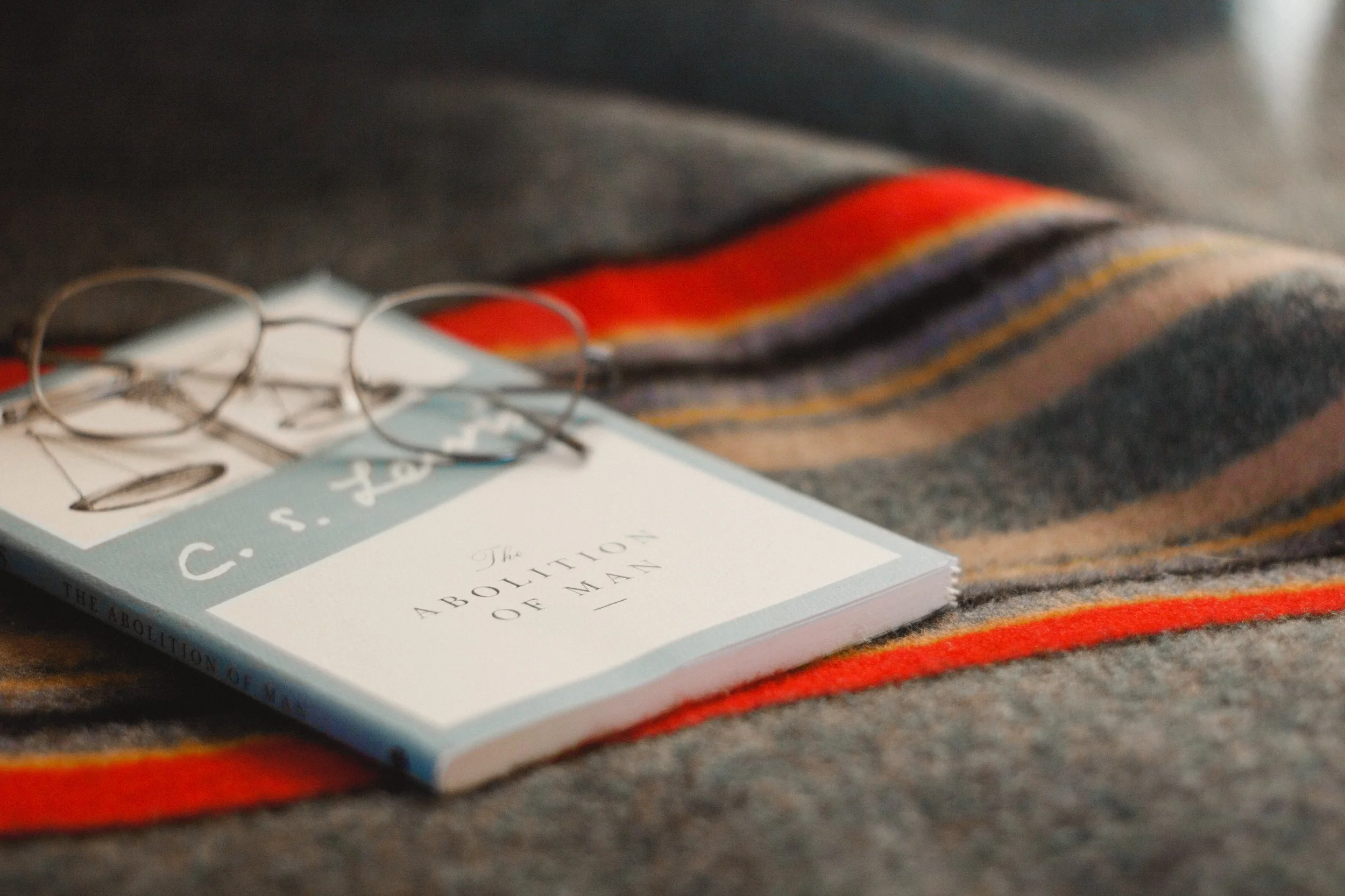 A book titled 'The Abolition of Man' by C.S. Lewis lies on a multicolored striped blanket with a pair of eyeglasses resting on top.