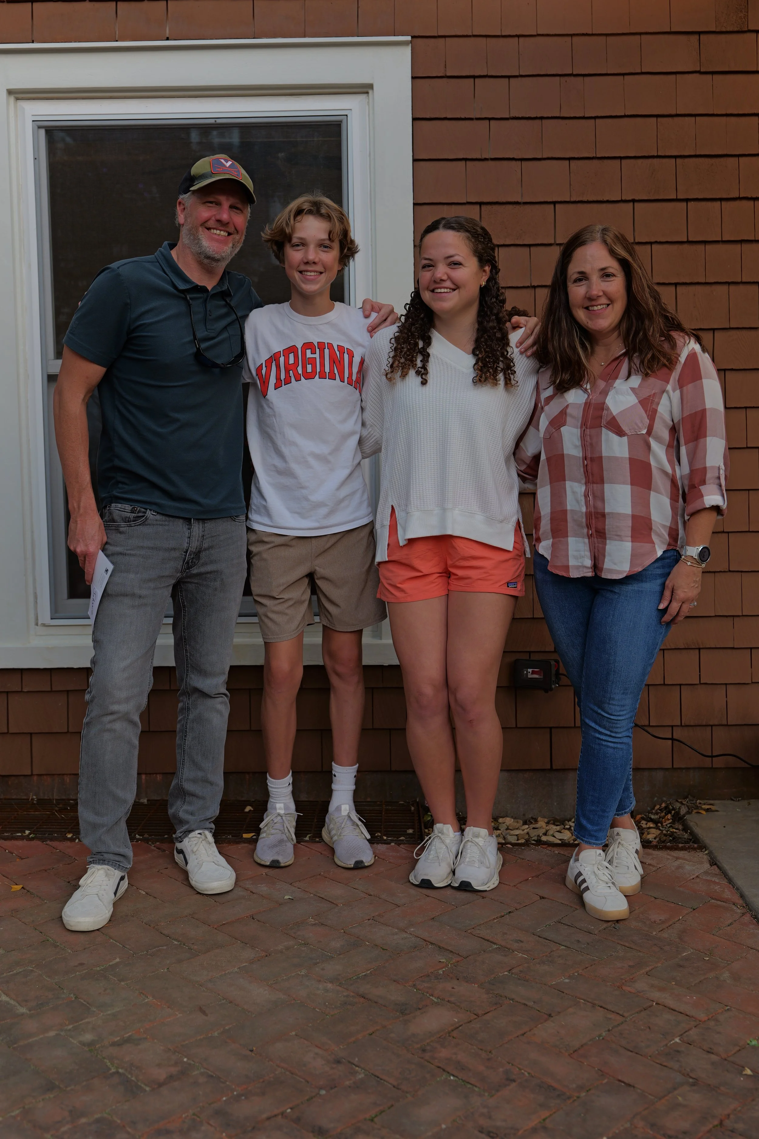 A group of four people standing outdoors, smiling, with a house wall and window behind them.
