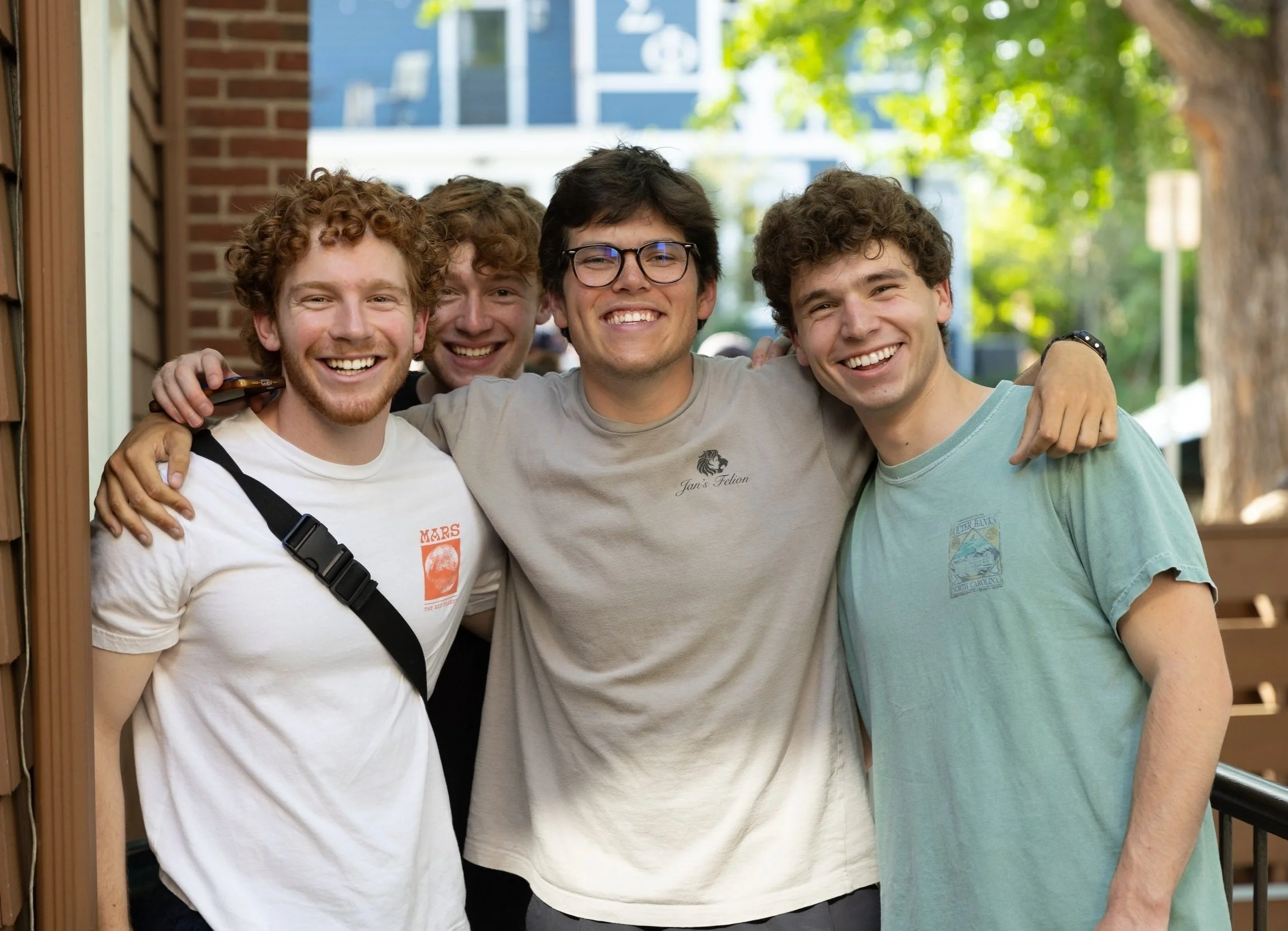 Group of five young men smiling and hugging outdoors on a sunny day.