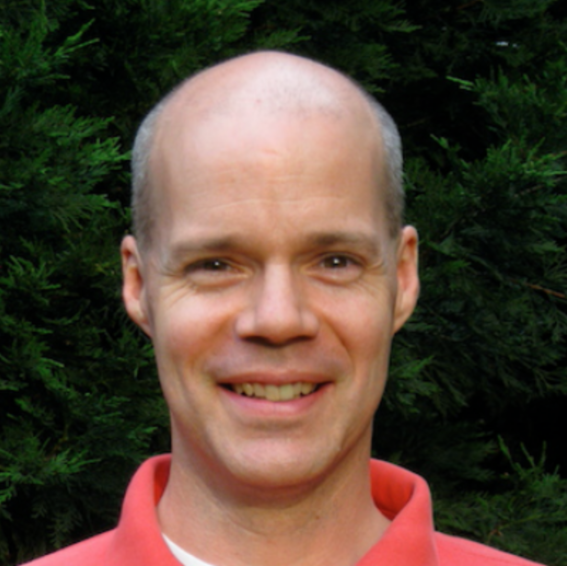 A smiling man with a bald head, wearing a red collared shirt, standing outdoors with green foliage in the background.