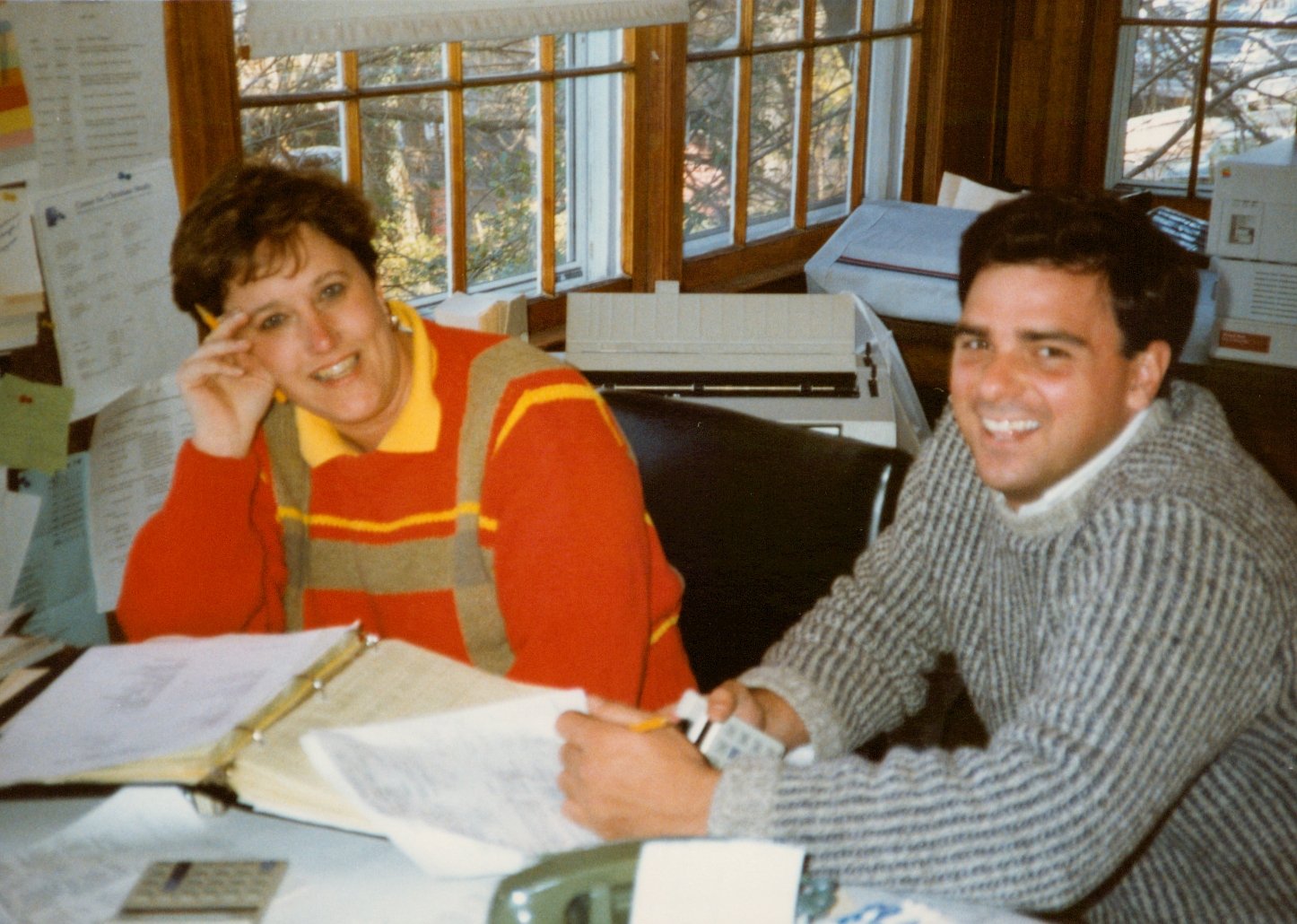 A woman and a man sitting at a cluttered office desk, smiling at the camera. The woman is wearing a red sweater with yellow and beige stripes, resting her head on her hand. The man is holding a phone and has dark hair, wearing a gray and white stripe