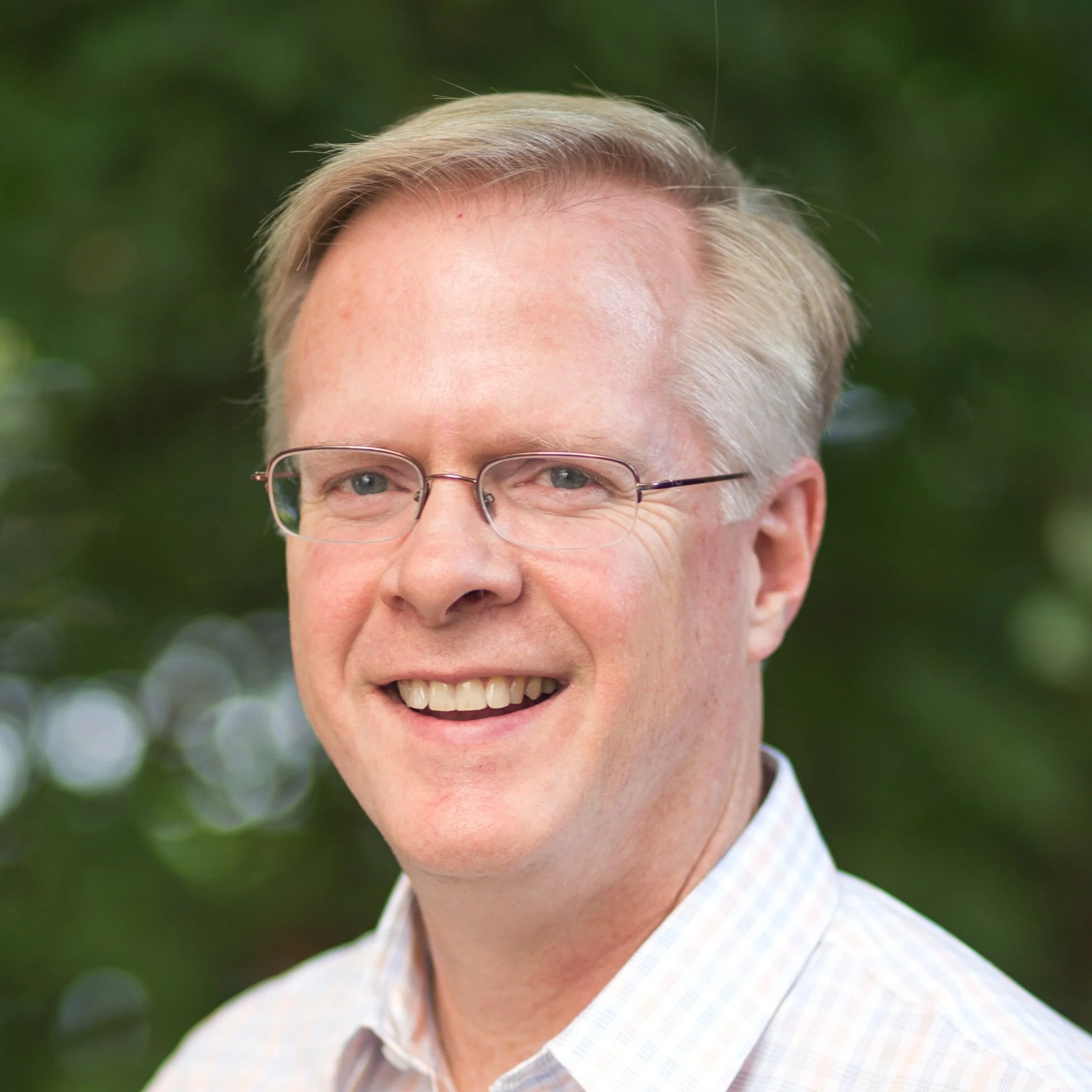 Portrait of a smiling man wearing glasses and a light checkered shirt, outdoors with a blurred green background.