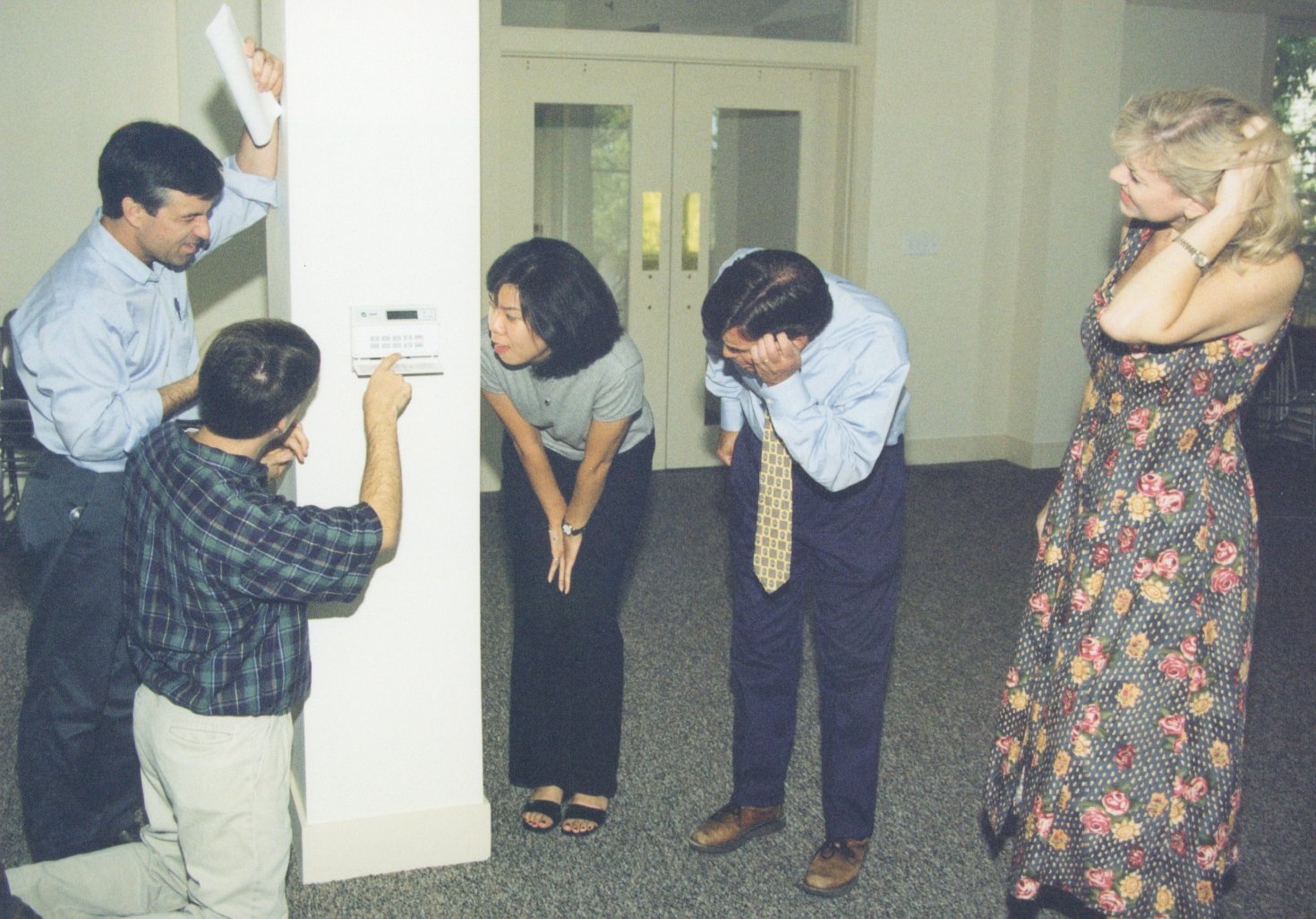 A group of six people gathered in an office, looking at an alarm system panel on the wall. One person is pointing at the panel while others react with concern or surprise.