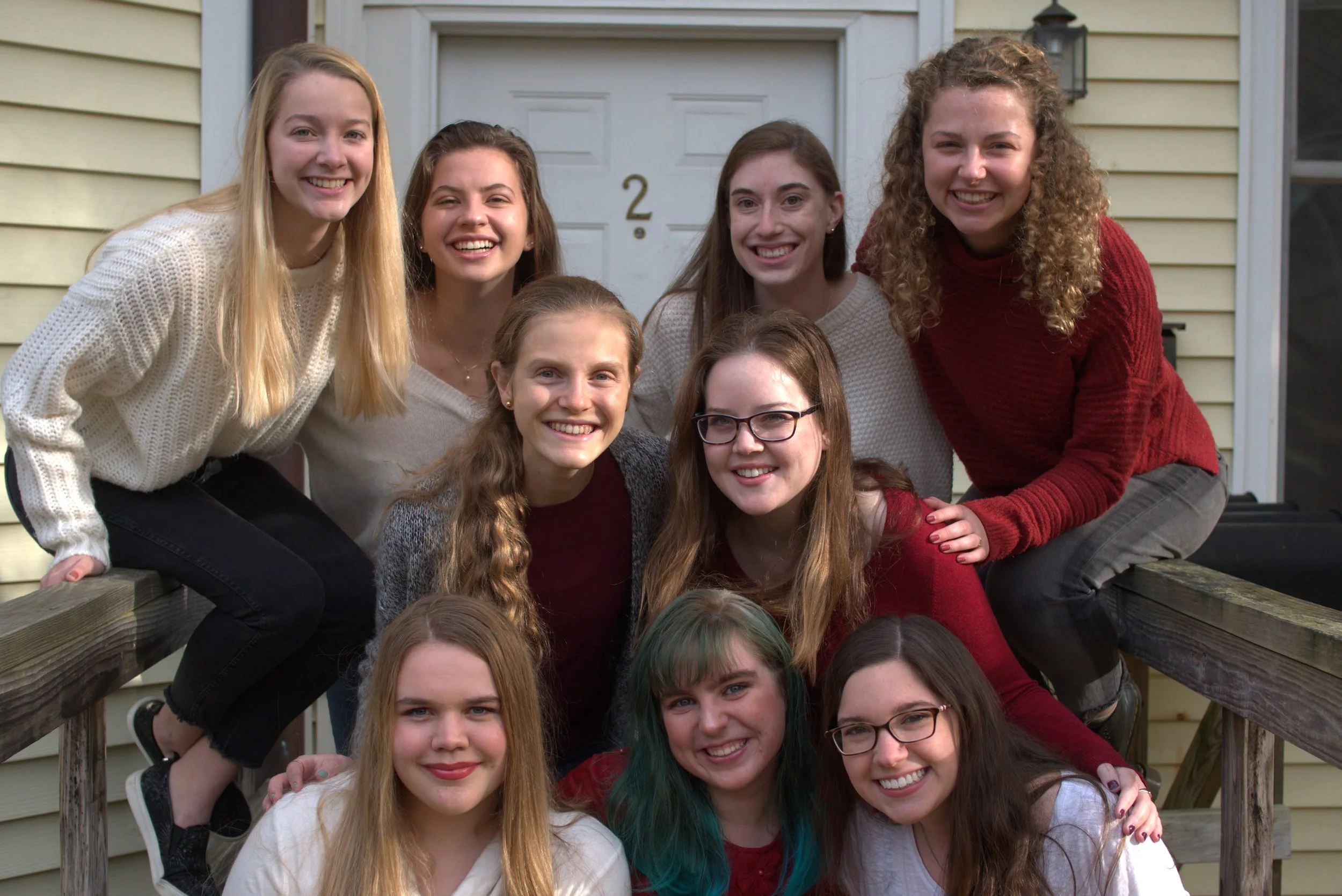 Group of ten young women posing on a wooden porch in front of a beige siding house, smiling at the camera.