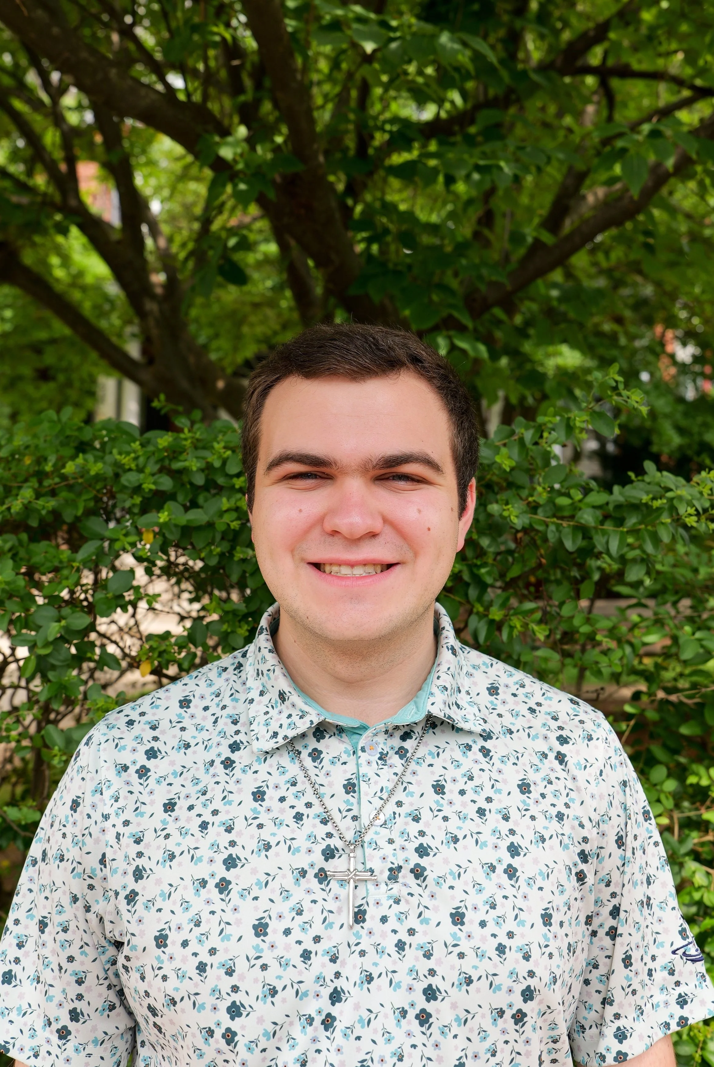 A young man with short dark hair smiling outdoors in front of green leafy trees and bushes, wearing a floral patterned short-sleeve shirt and a necklace with a cross pendant.