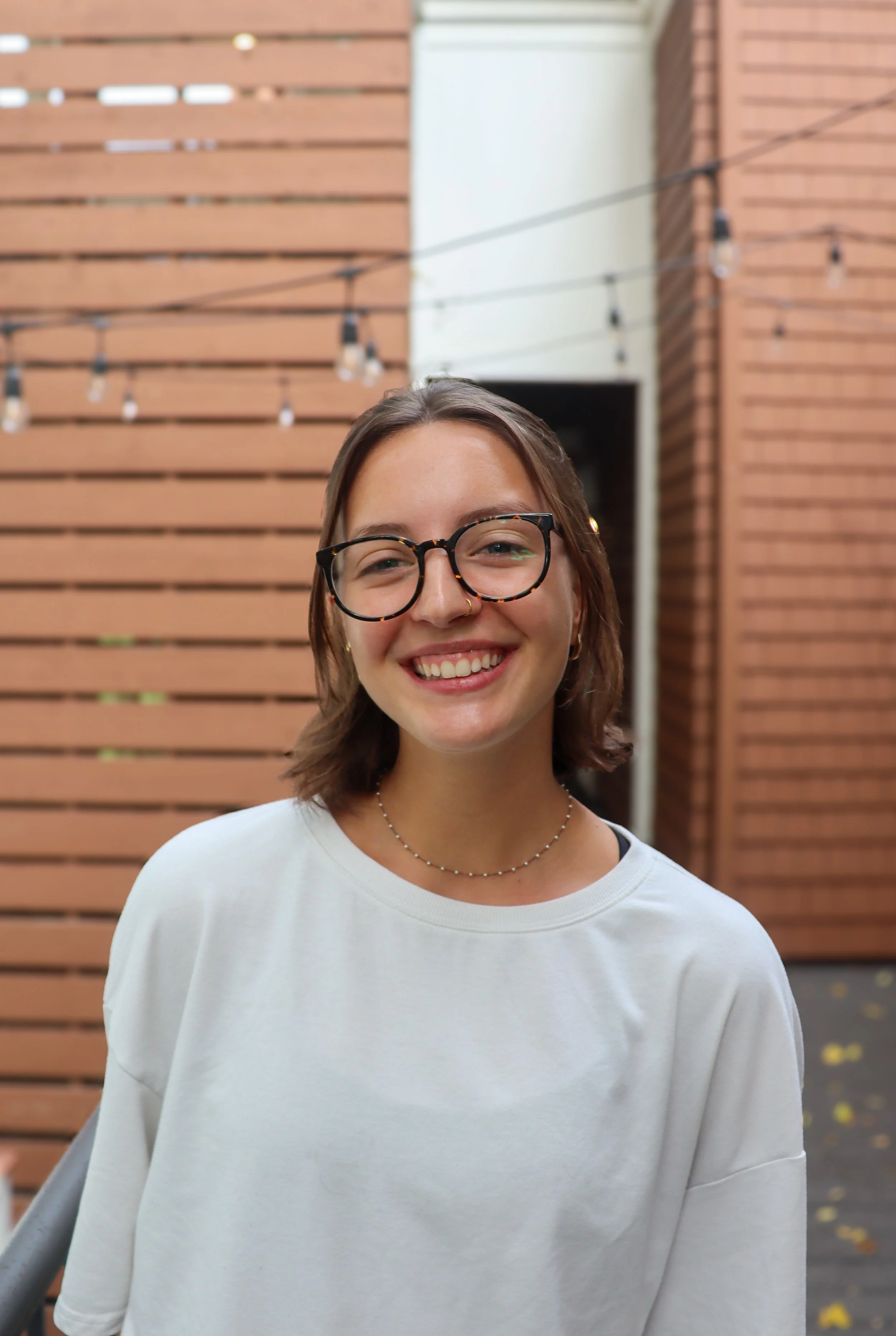 Close-up of a smiling woman with blue eyes, short brown hair, wearing gold hoop earrings, a small nose ring, a thin gold necklace, and a gray and white striped shirt, standing outdoors in front of a wooden structure and a building with string lights.