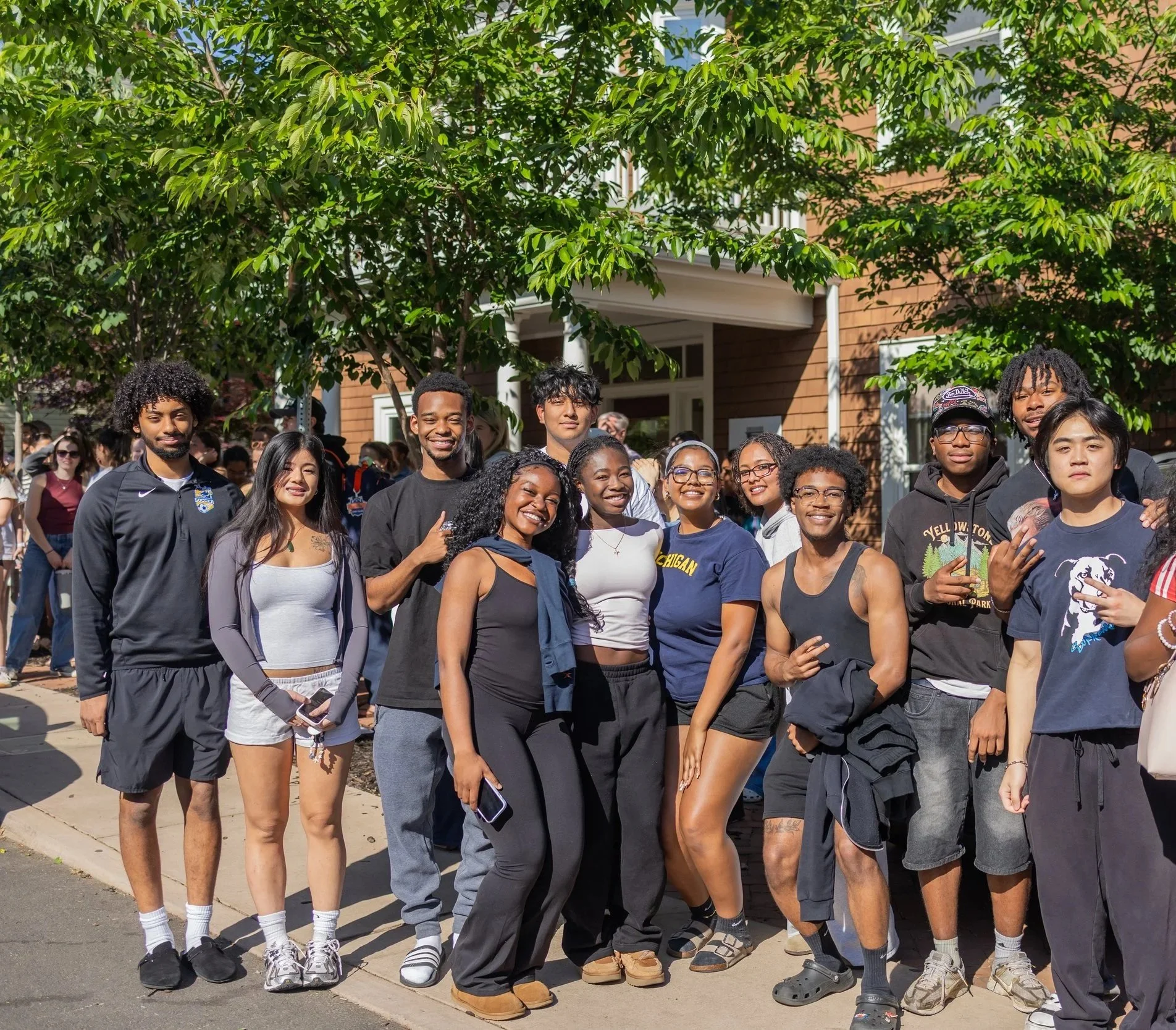 A diverse group of young people smiling and standing together outdoors in front of a tree and a house with a wooden exterior, during daytime.