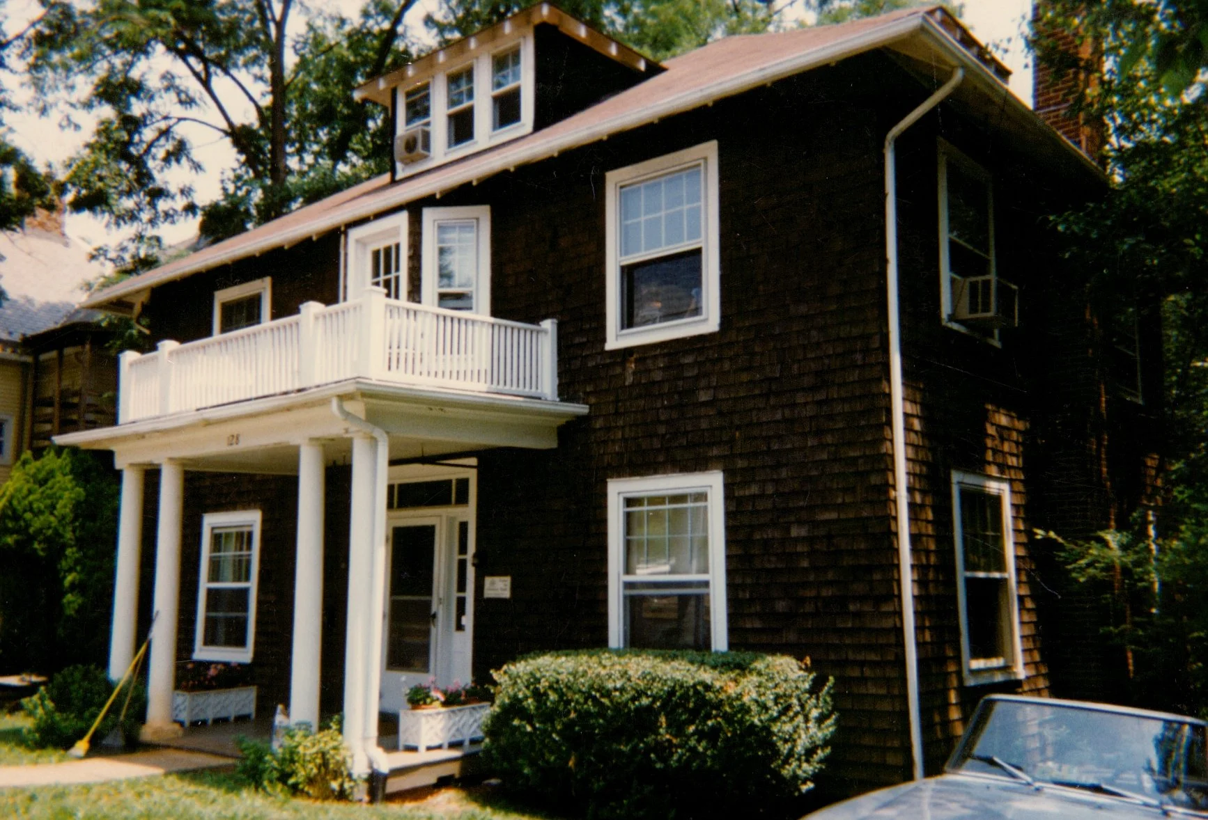 A multi-story house with brown wooden shingles, multiple white-framed windows, a stairway leading to a side entrance, and a car parked on a driveway in the background, surrounded by trees and greenery.