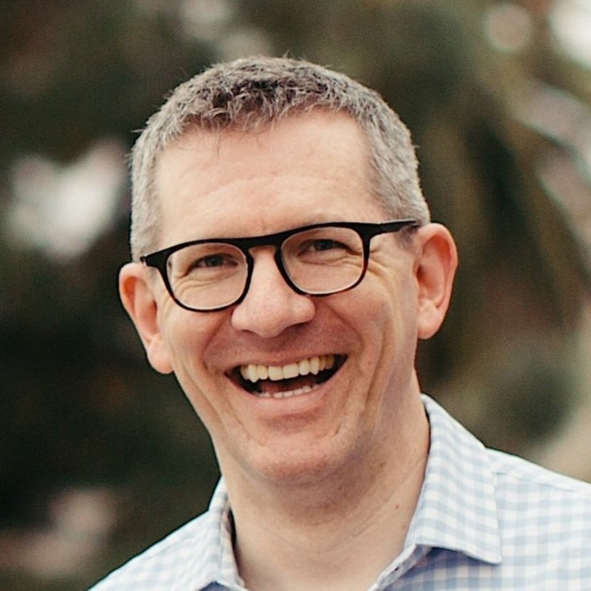 A man with short gray hair, wearing black glasses and a light-colored checkered shirt, smiling outdoors.