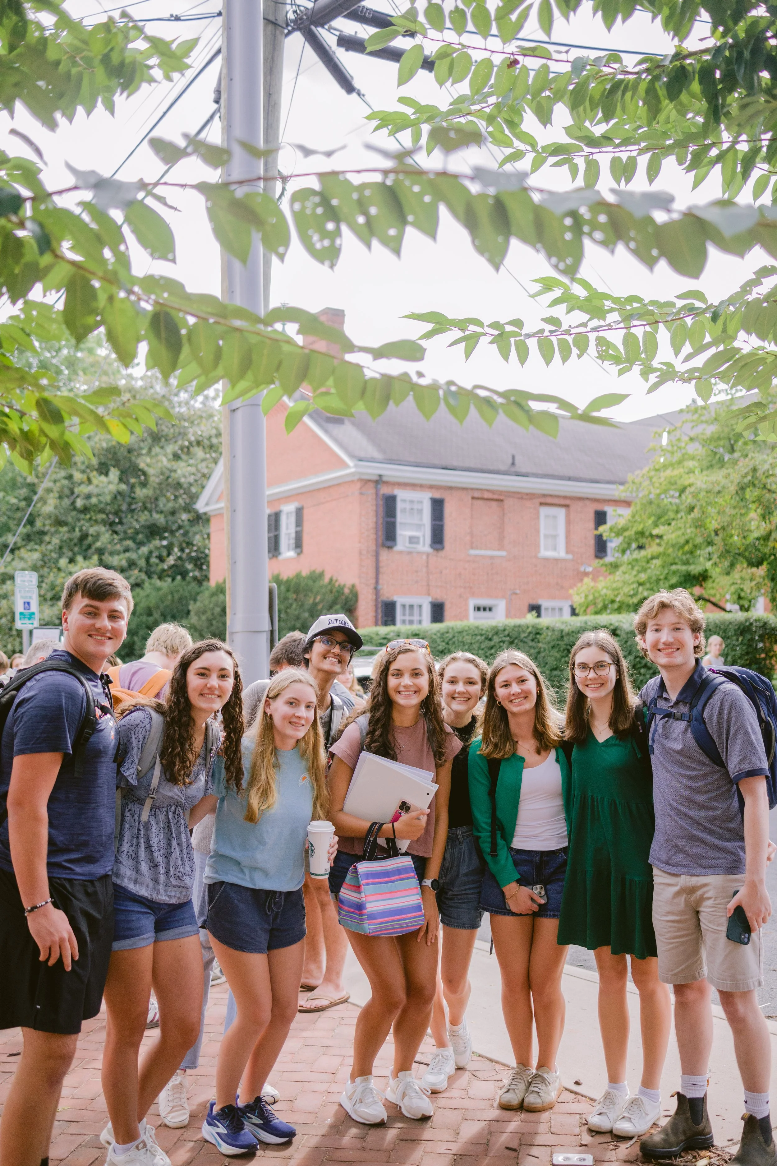Group of young people standing outdoors on a brick sidewalk, smiling, with trees and a brick house in the background.