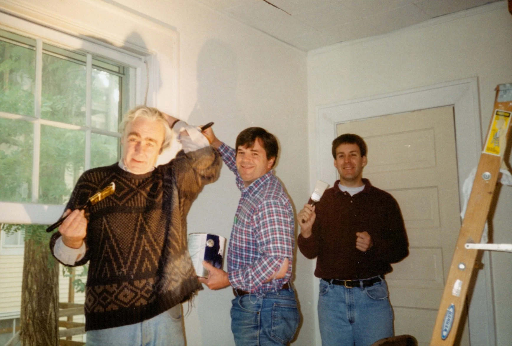 Three men painting a wall inside a house with a ladder and a window in the background.
