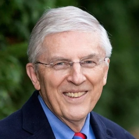 Portrait of a smiling older man with glasses, gray hair, in a navy jacket, blue shirt, and red tie, outdoors with green foliage background.