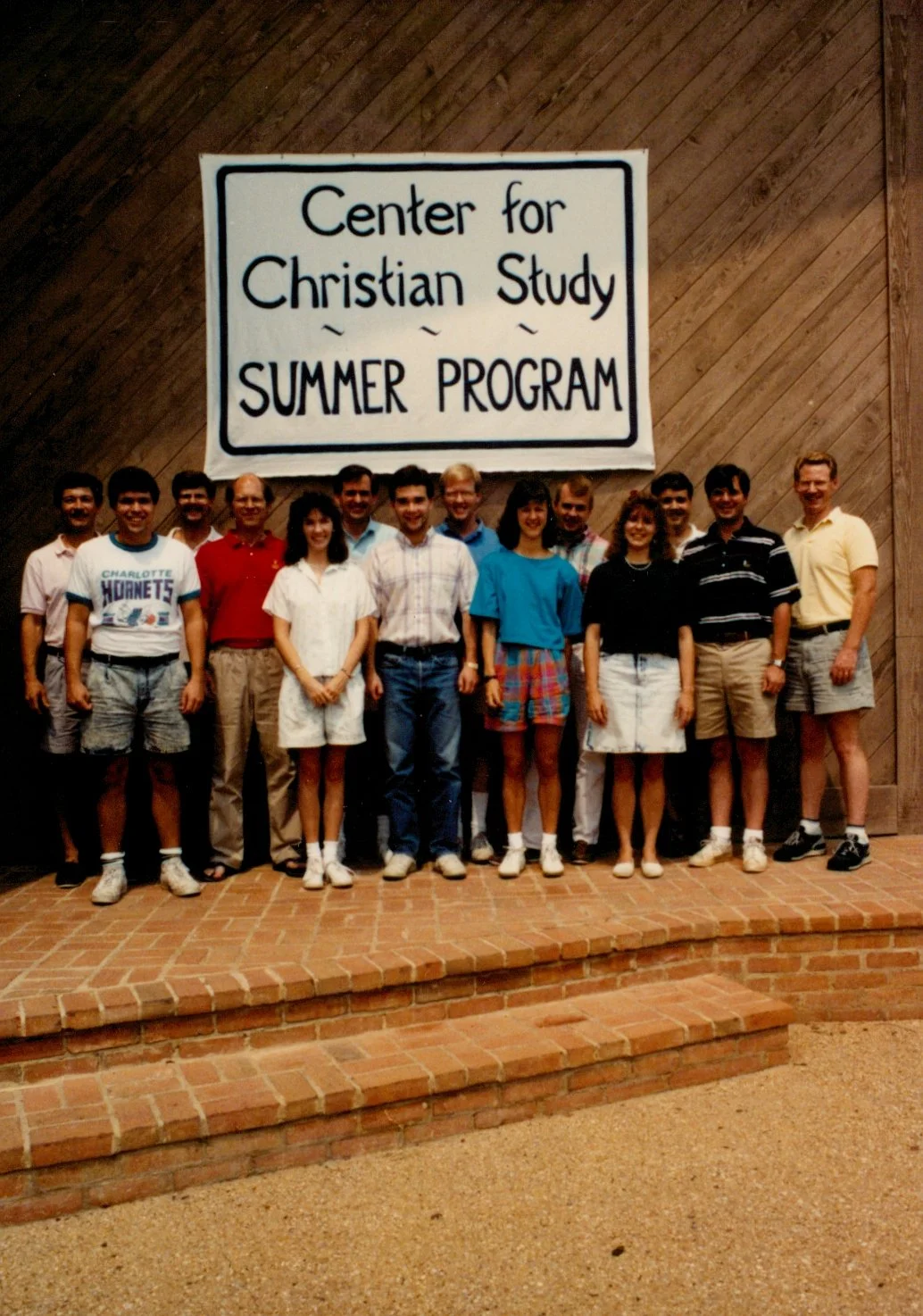 Group of people standing in front of a sign that reads 'Center for Christian Study Summer Program,' on a brick platform with a wooden wall background.