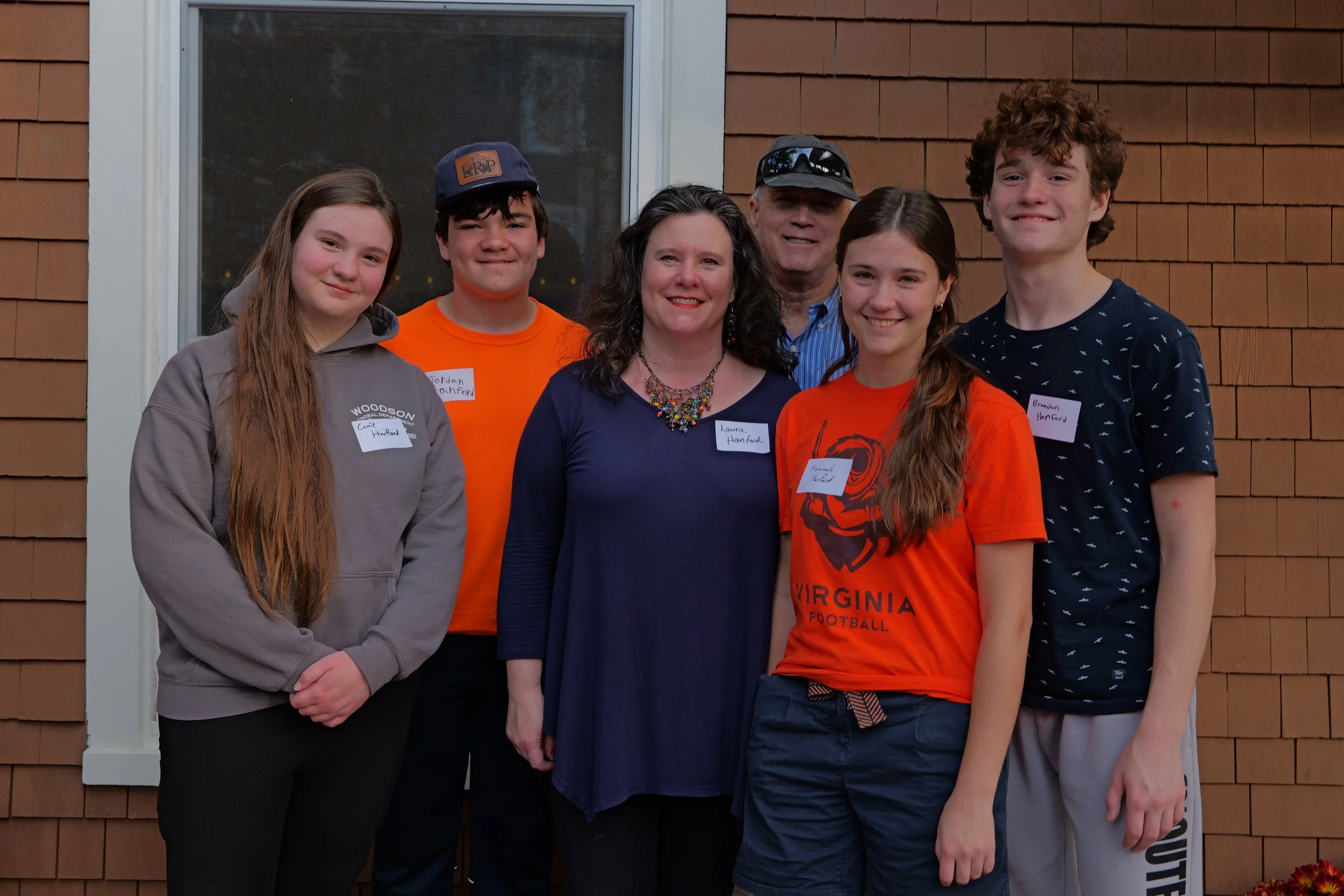 Group photo of six people standing in front of a brick wall, including three teenagers with name tags and three adults, smiling for the camera.