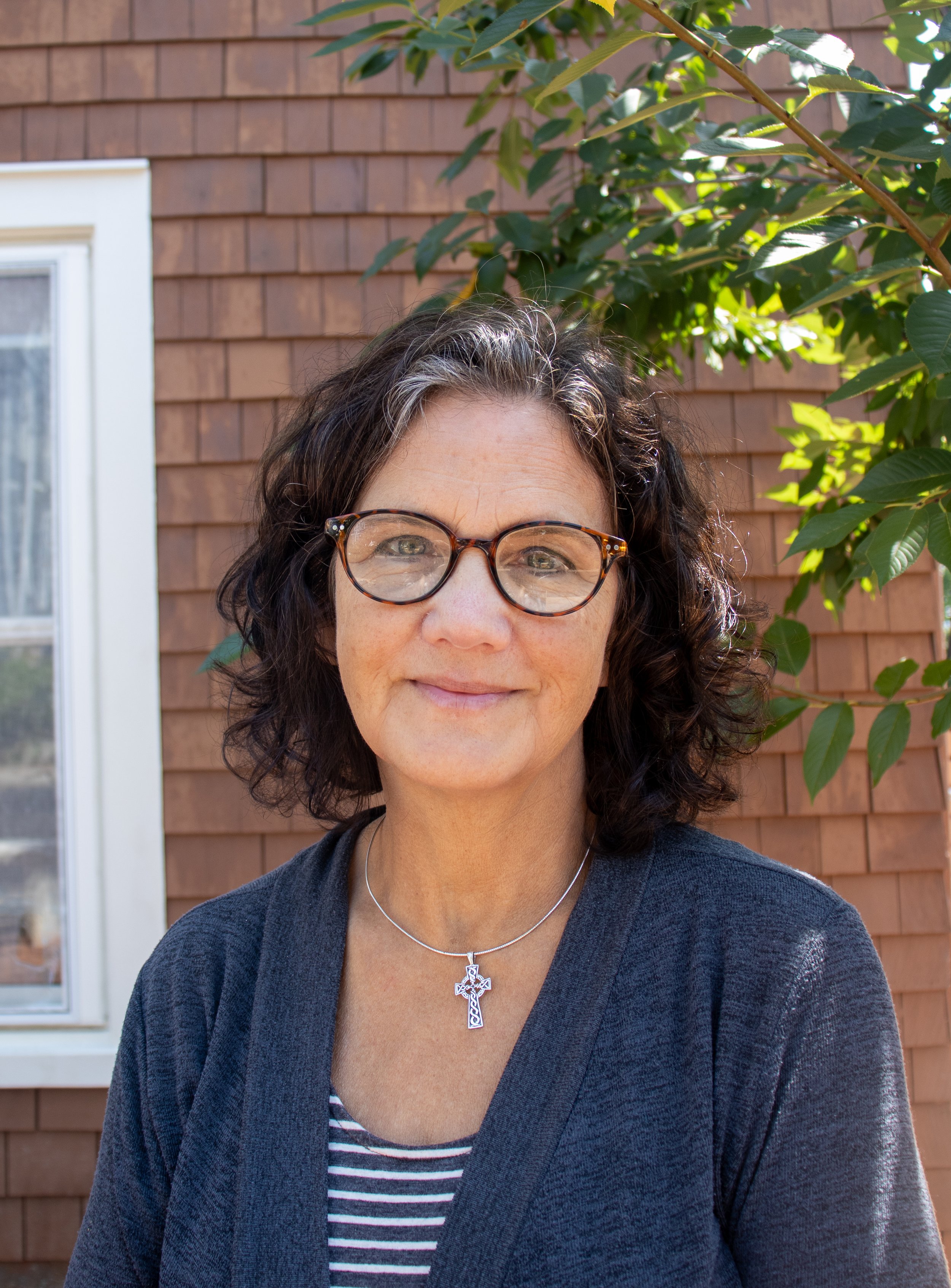 A woman with curly dark hair and light eyes smiling outdoors in front of a brown wooden house and a leafy green plant, wearing a navy cardigan, striped shirt, and a silver cross necklace.