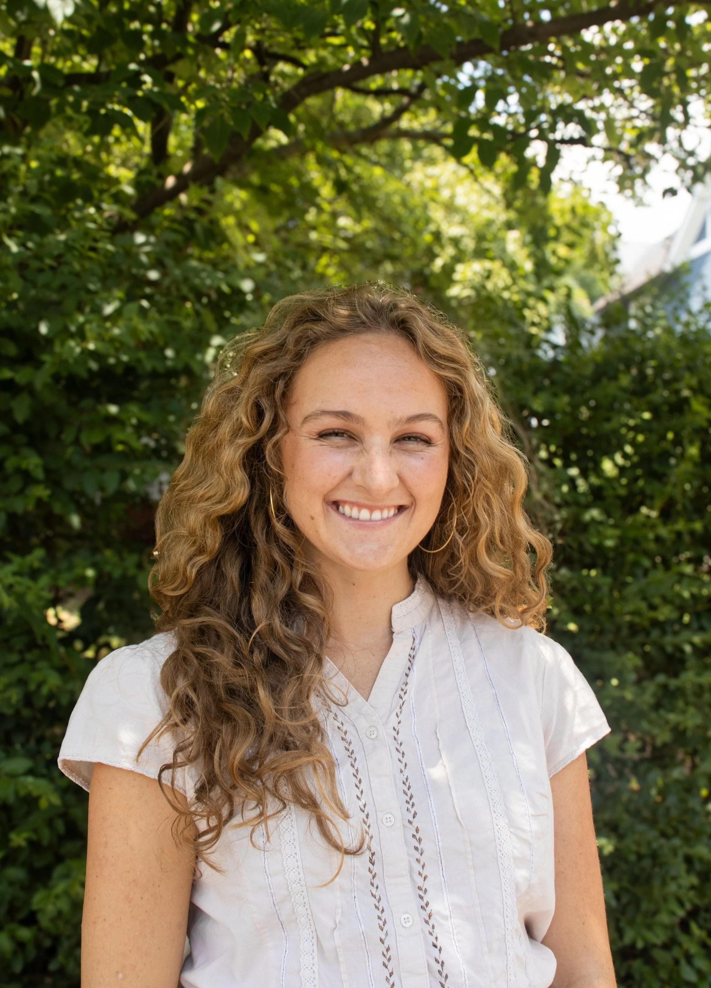A woman with curly blonde hair smiling outdoors with green trees in the background.