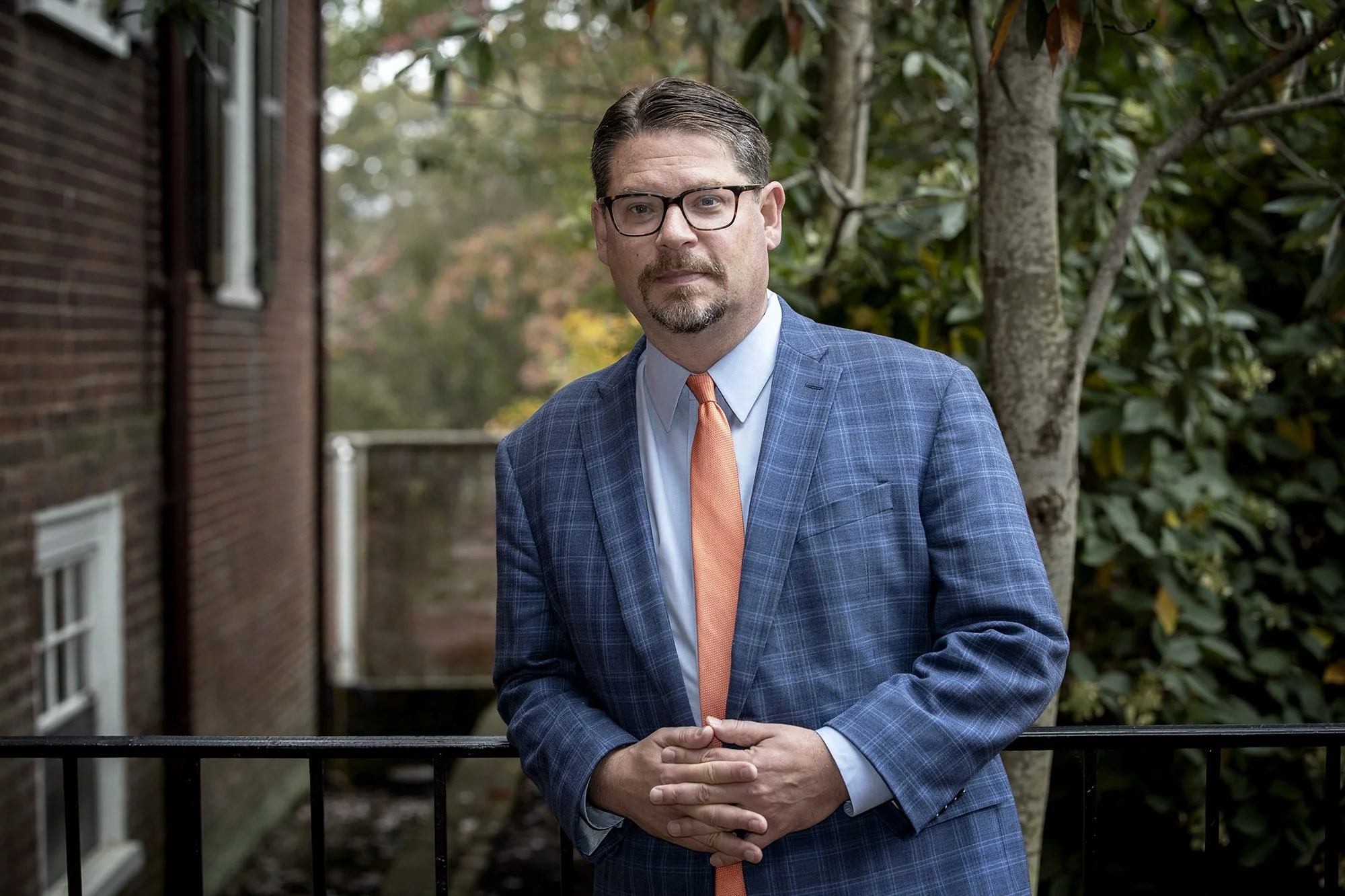 A man with glasses, a beard, and dressed in a blue plaid suit with a white shirt and orange tie, standing outdoors near a tree and a brick building.