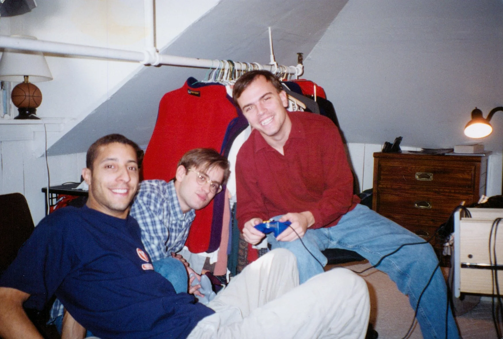 Three young men with short hair and glasses, smiling and sitting in a room with a sloped ceiling. One is holding a video game controller. Behind them are clothes hanging on a rack and a wooden dresser.