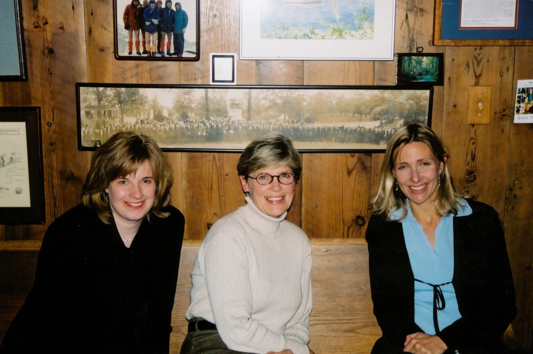 Three women sitting side by side, smiling, in front of a wooden wall decorated with framed photos and artwork.