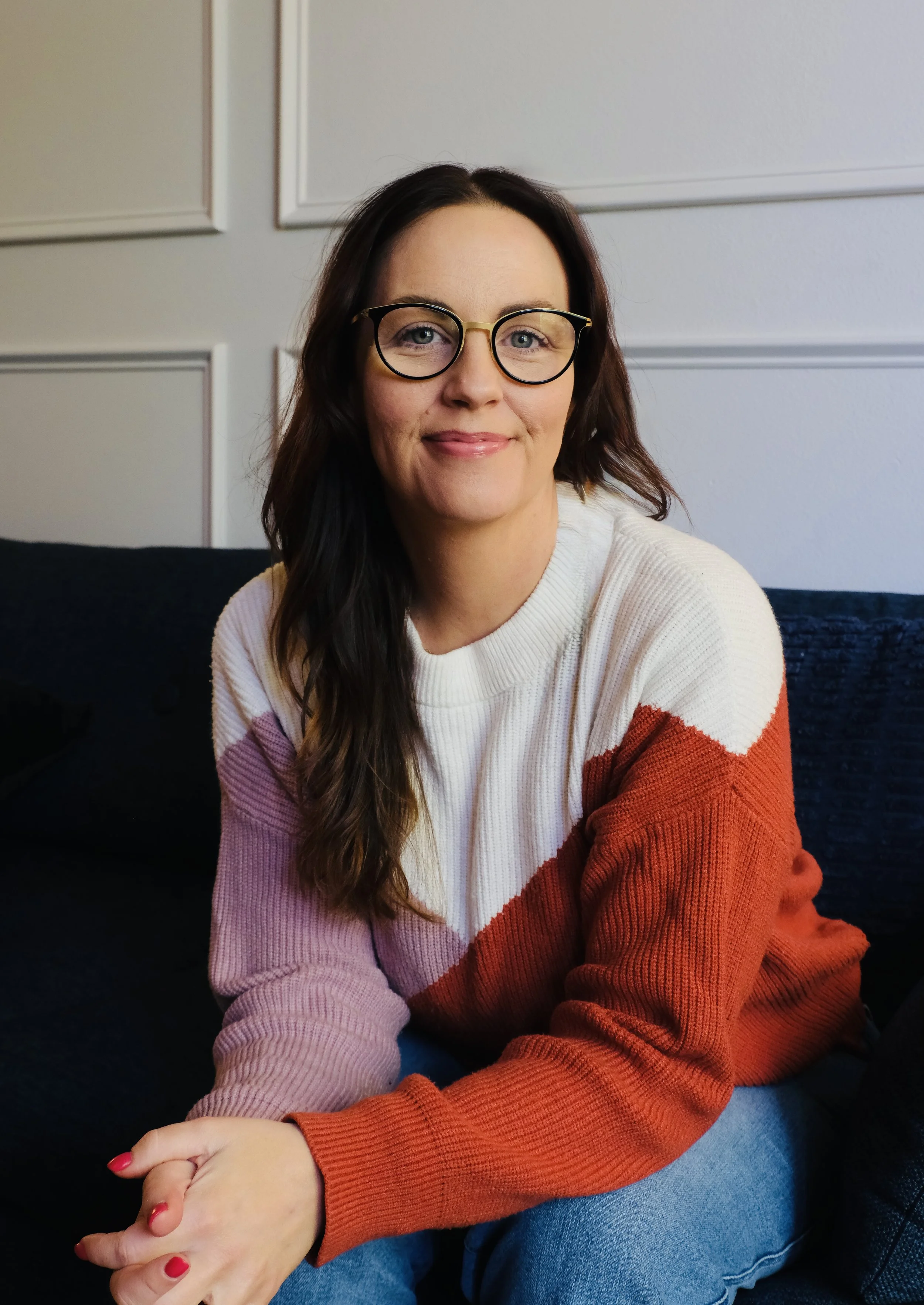 A woman with long dark hair and glasses smiling, wearing a multicolored sweater, sitting on a dark blue couch with a light-colored wall in the background.