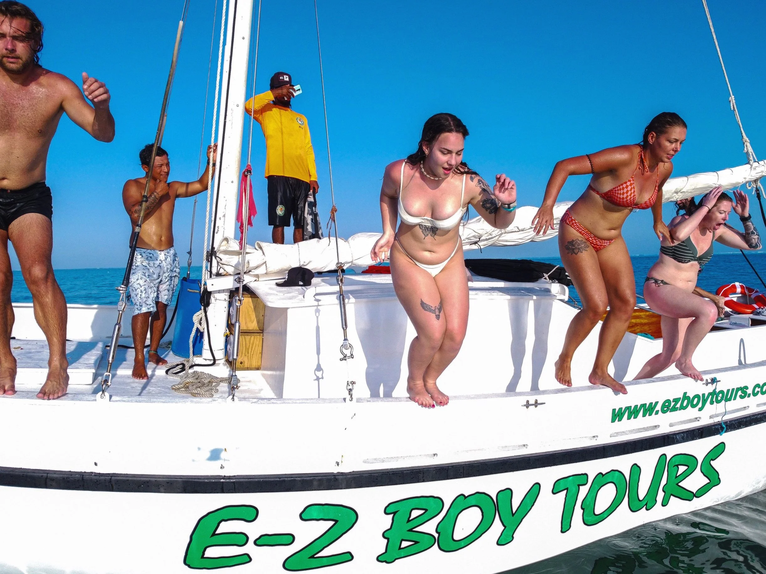 Group of people jumping off an E-Z Boy Tours sailboat into the water on a sunny day, with bright blue sky, some wearing swimsuits and some in casual summer clothing.