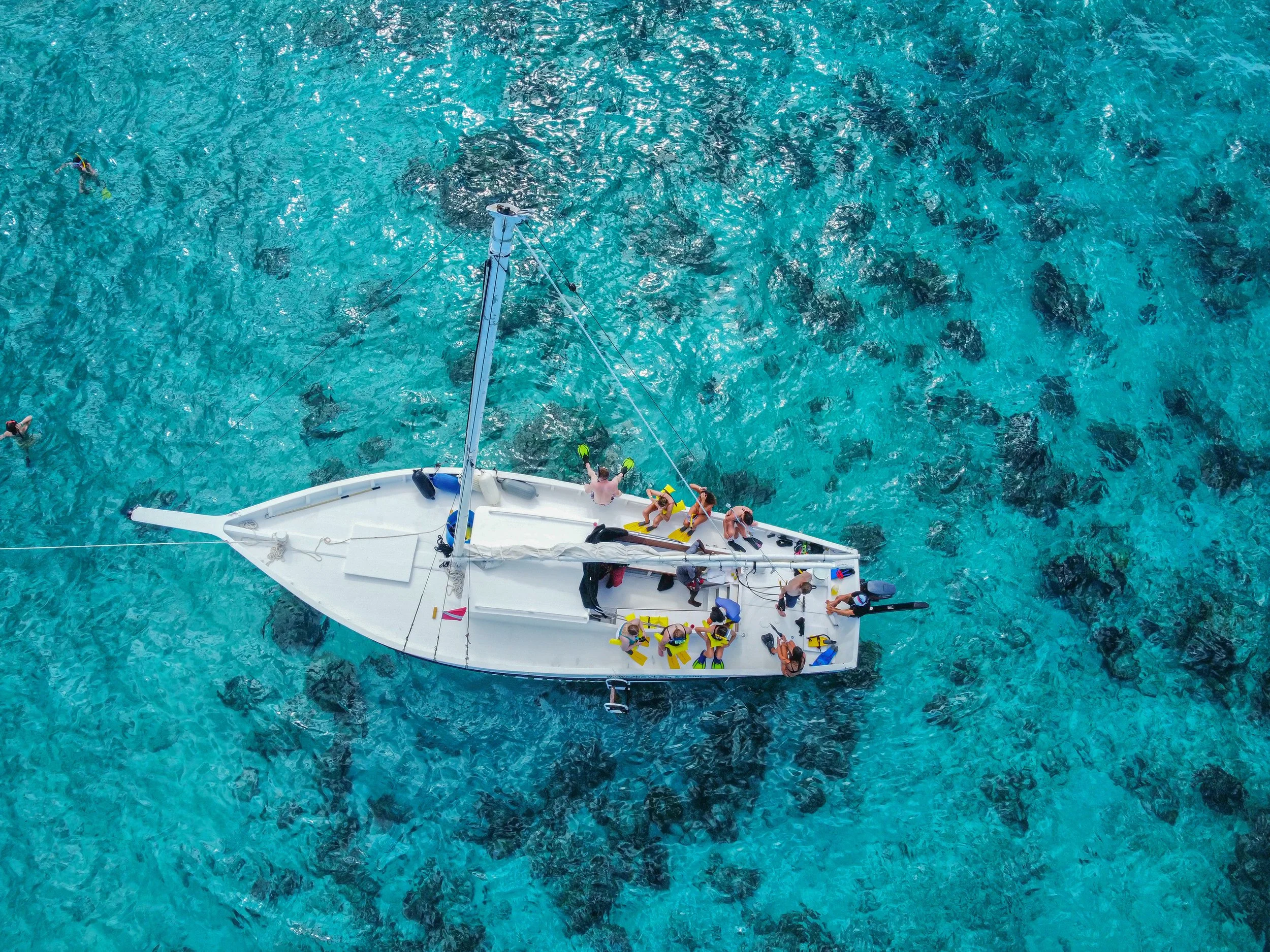 An aerial view of a white sailboat with a group of people on board, floating on clear blue water with visible dark patches indicating seaweed or rocks beneath.