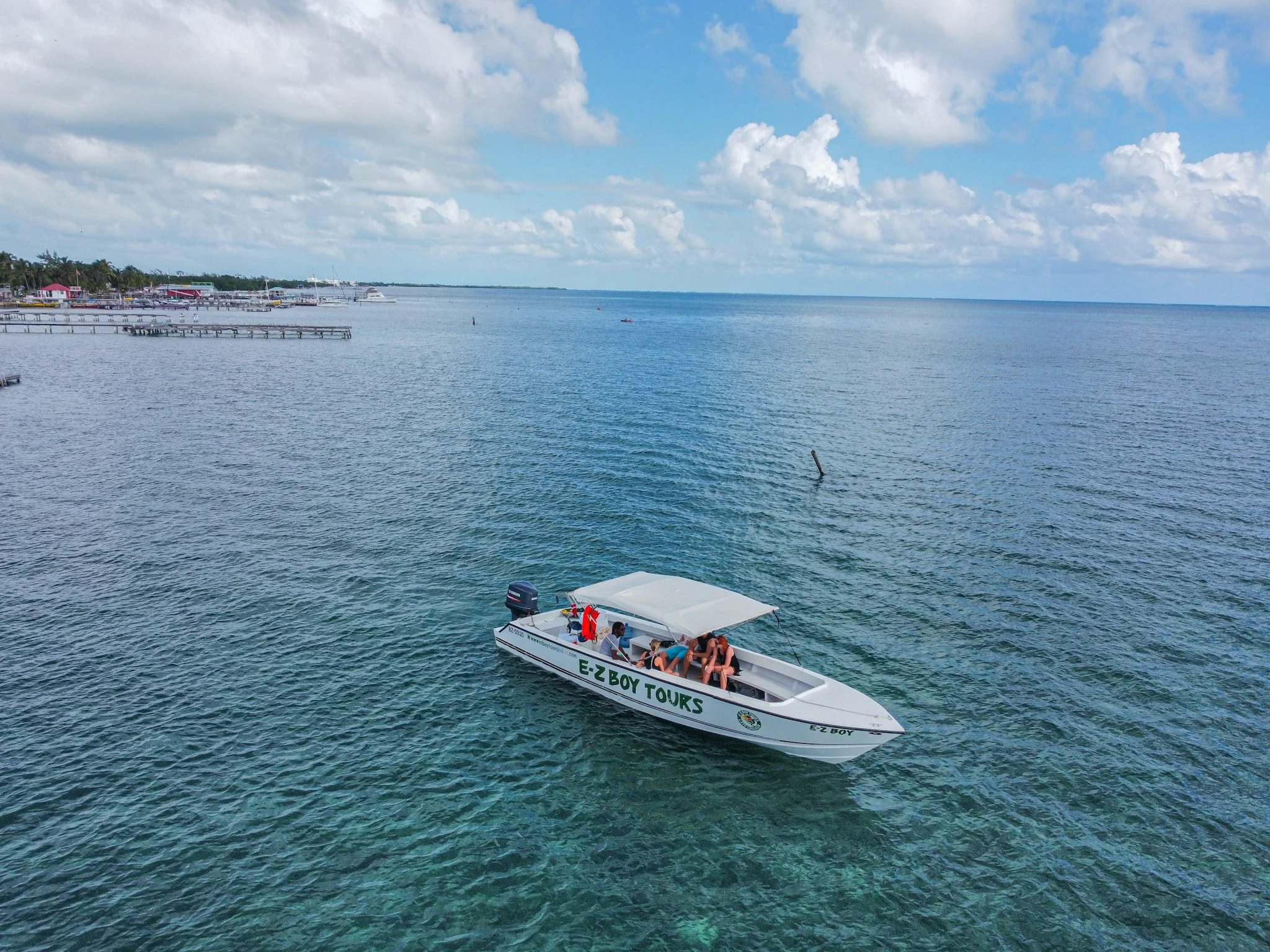 A boat with the text 'E-Z Boy Tours' is floating on calm ocean waters on a sunny day with partly cloudy skies; a marina with boats and buildings is visible in the background.