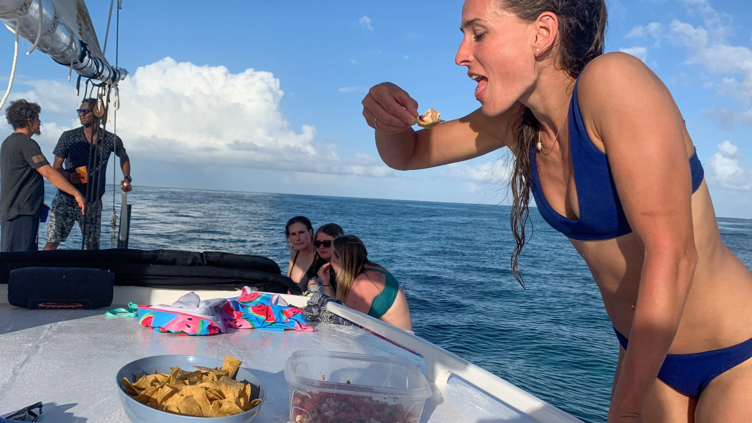 Woman in blue bikini eating on a boat with four other women sitting in the background, ocean and blue sky with clouds