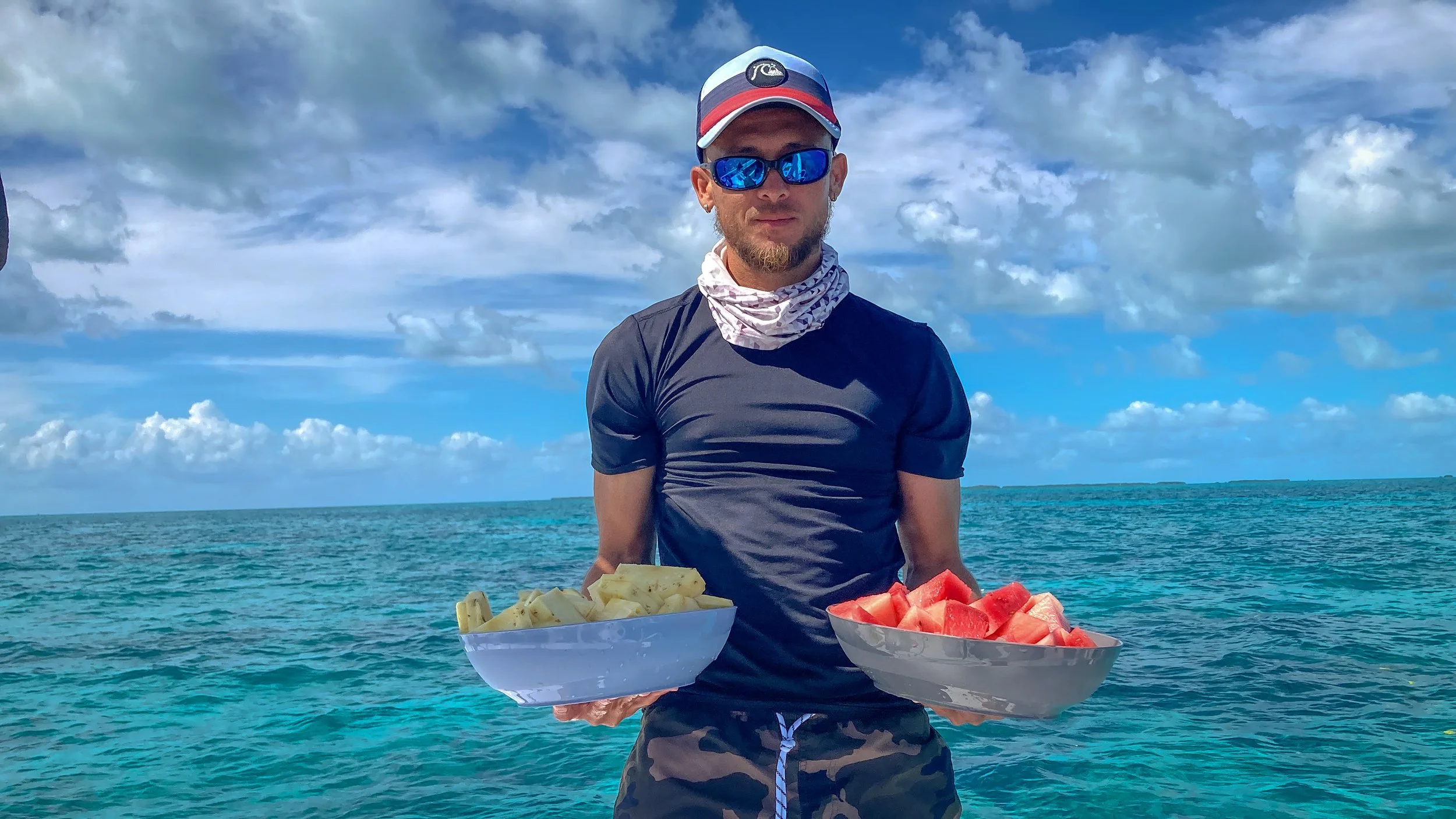 Man with sunglasses, hat, and neck buff holding two bowls of sliced watermelon and pineapple in front of ocean with cloudy sky.