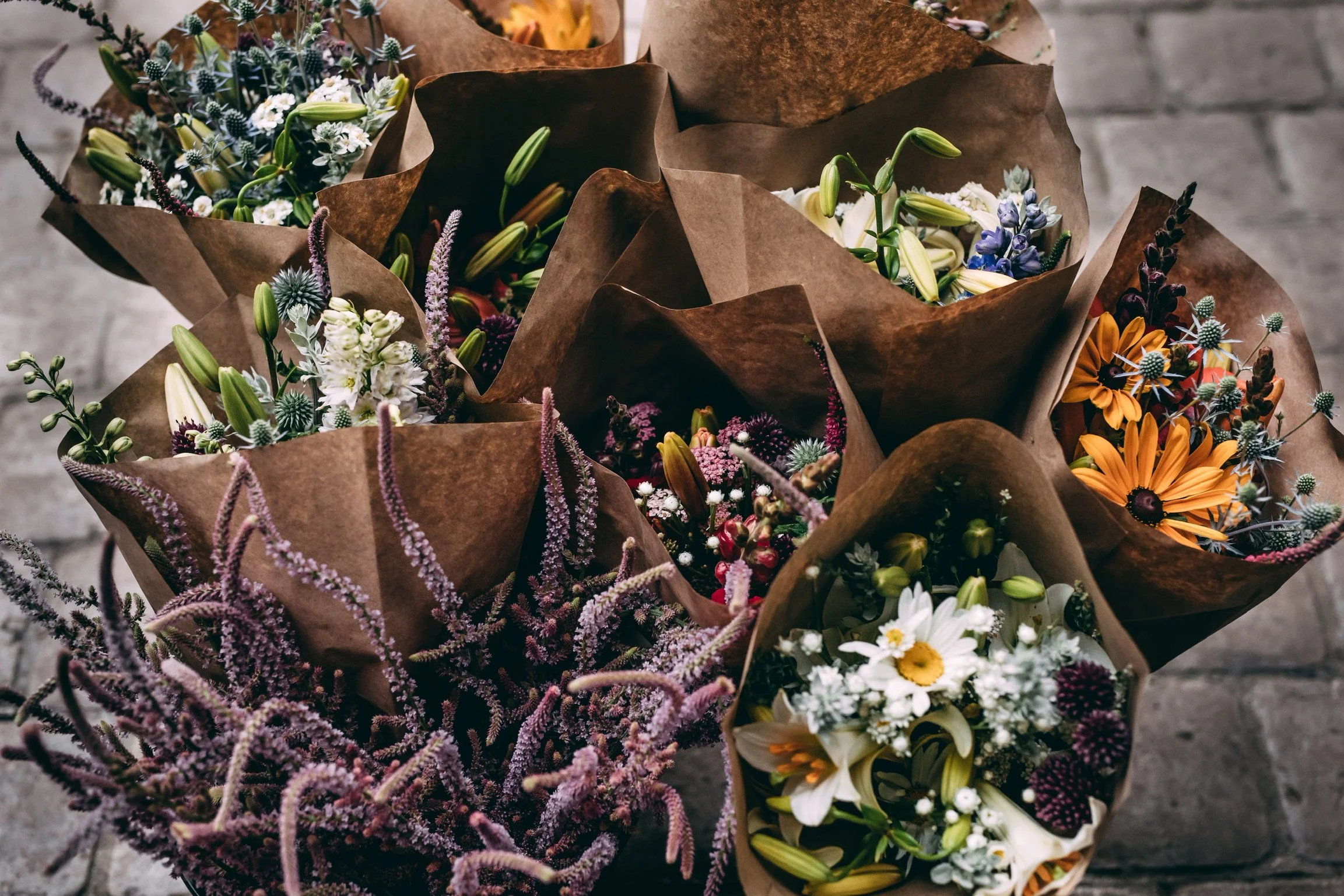 Multiple bouquets of flowers wrapped in brown paper on a concrete surface.