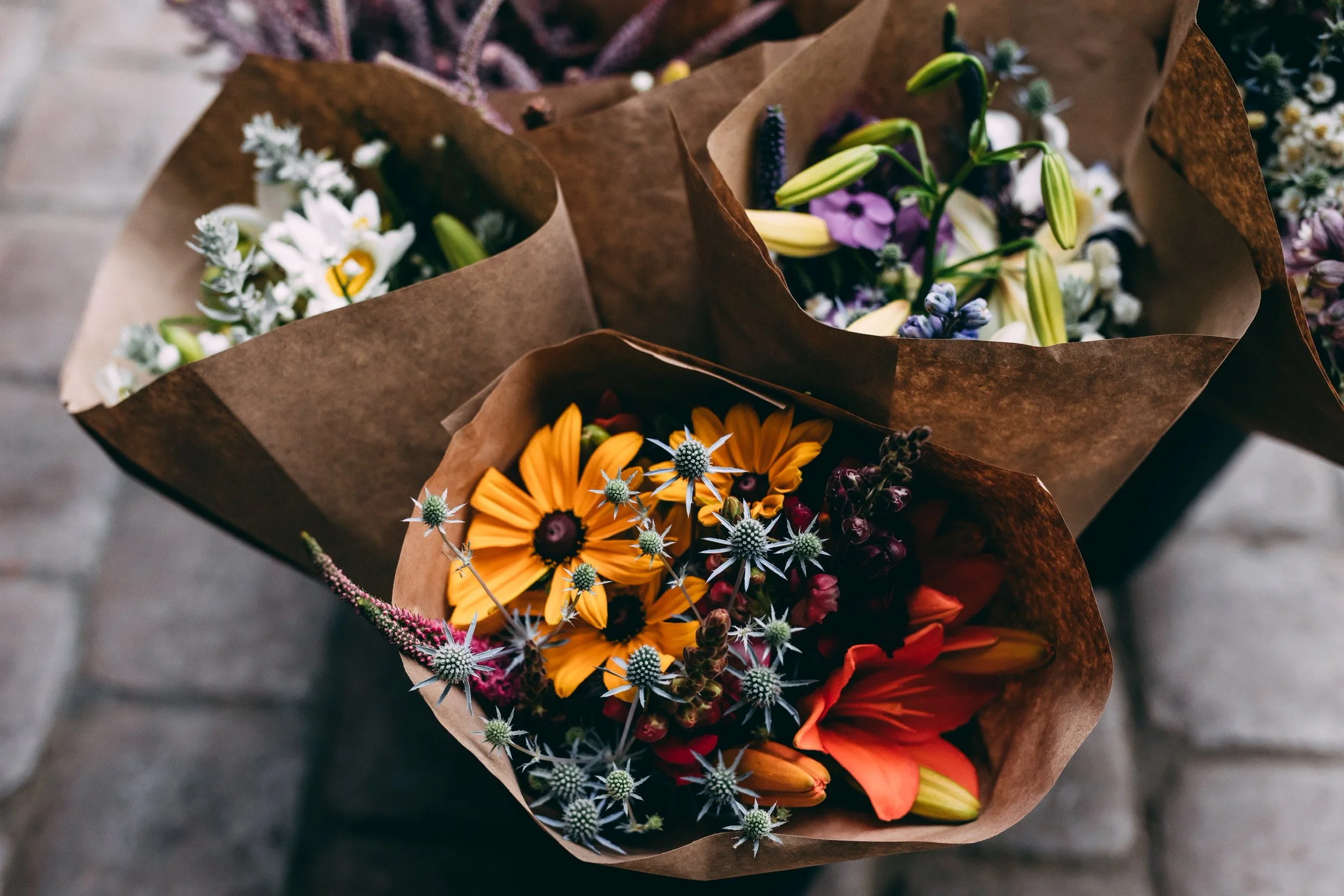 Colorful flower bouquets wrapped in brown paper, including orange, white, purple, and yellow flowers, on a wooden surface.