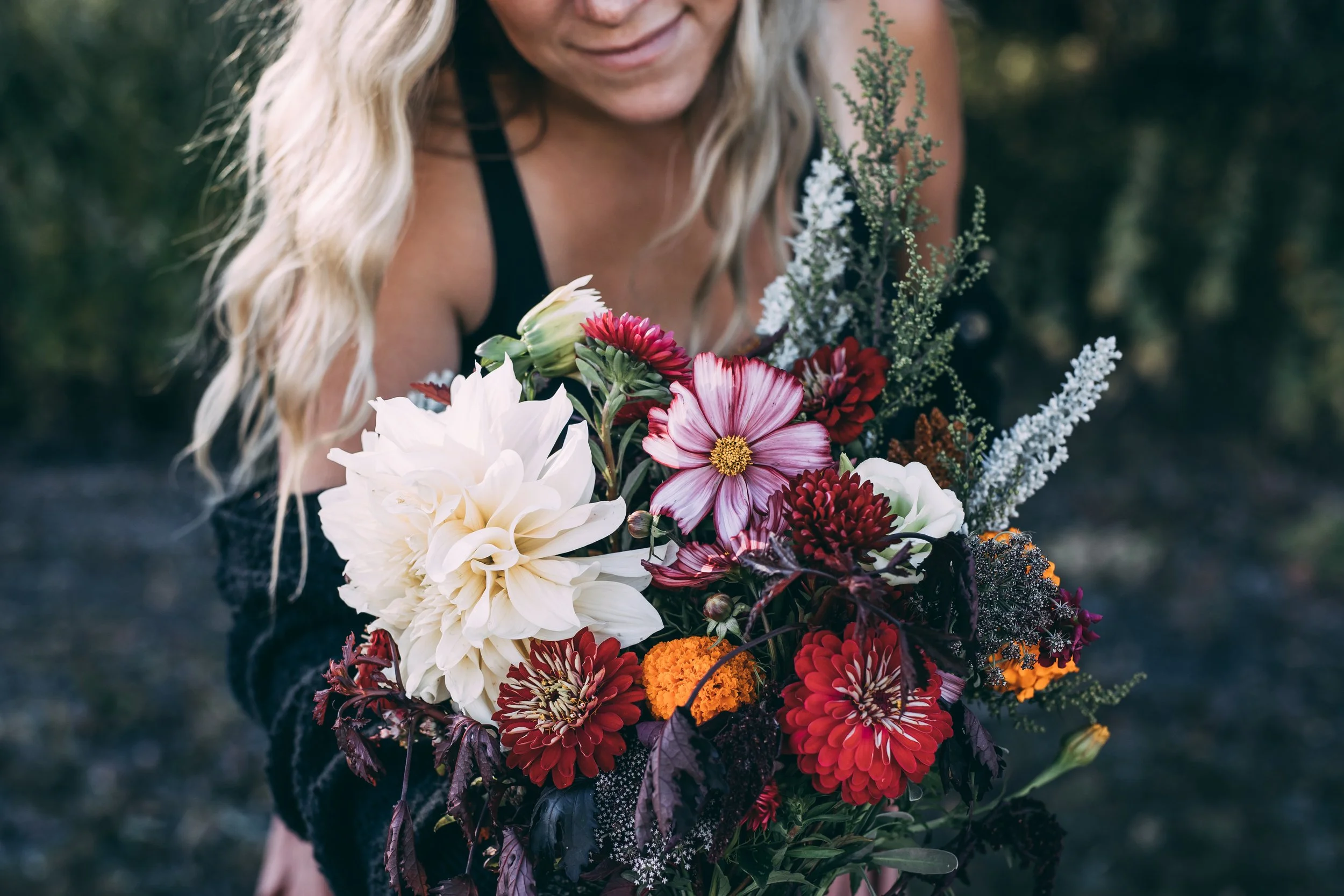 A person with blonde curly hair holding a colorful bouquet of various flowers including white, red, pink, purple, and orange blossoms with greenery, outdoors during daylight.