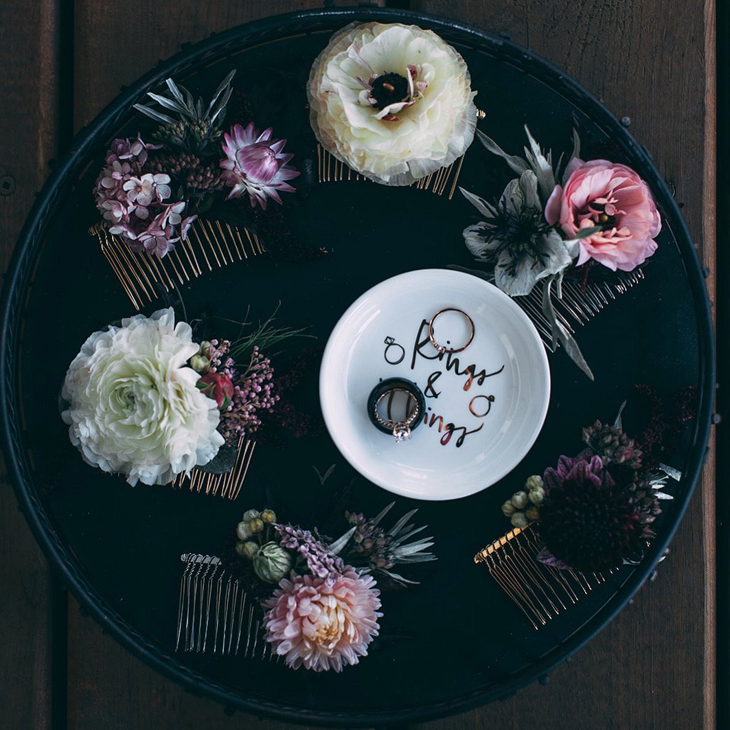 A black round tray decorated with pink, white, and purple flowers and hair combs, with a white dish in the center holding two rings, and the words "Mrs & Mr" written inside the dish.