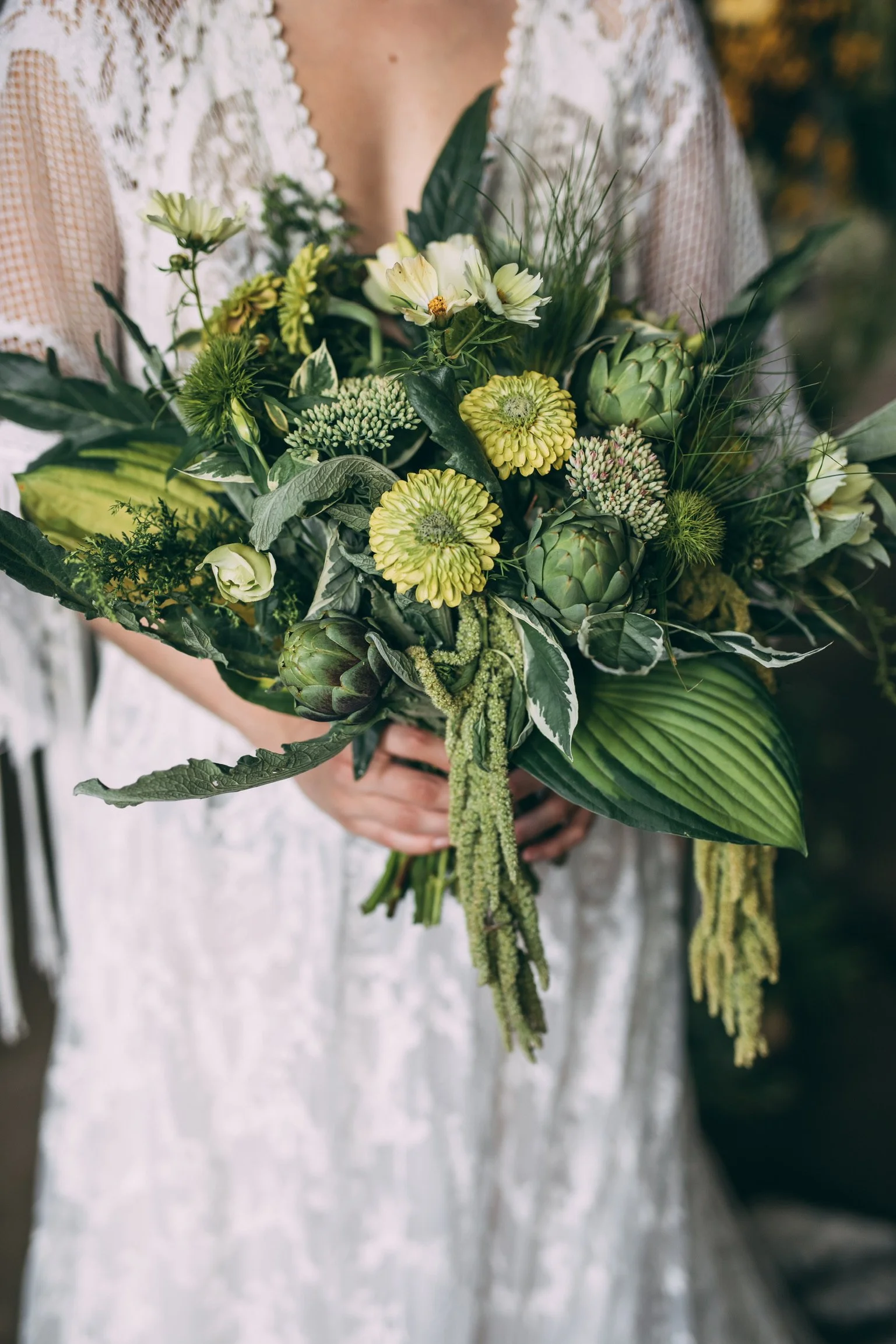 Person holding a bouquet of green and white flowers and greenery, wearing a white dress with lace details.